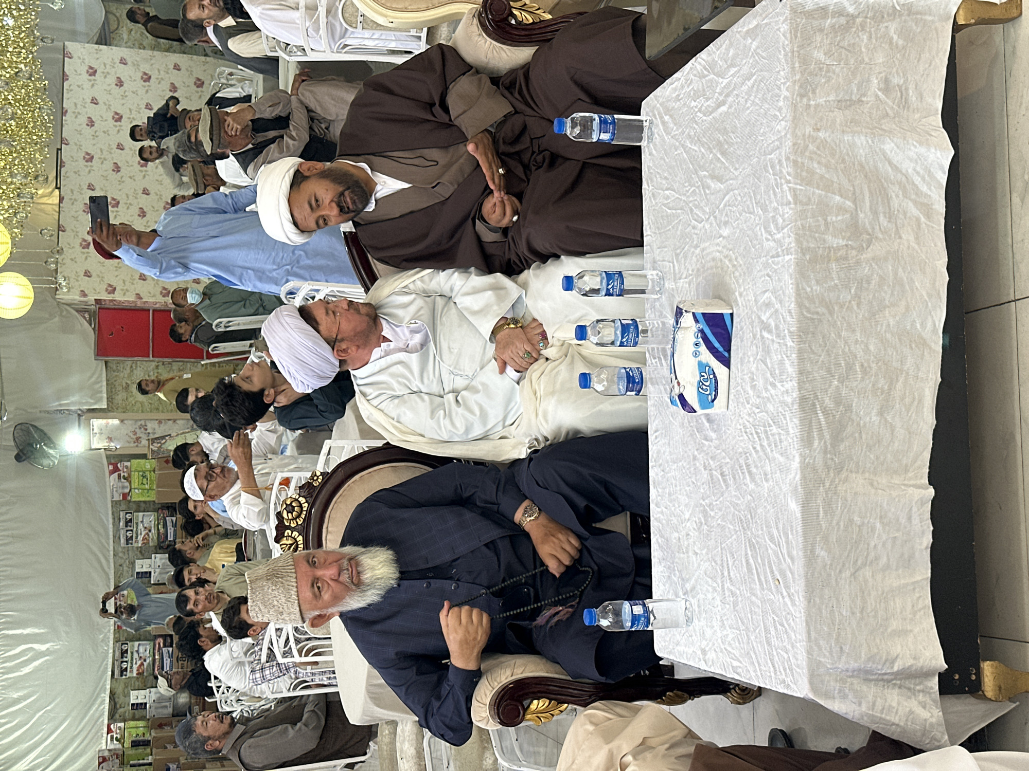 **Scene:** An indoor gathering in Quetta City Tehsil, Pakistan.
**People:** A group of men are seated in rows. Three men sit closest to the table, adorned in traditional Islamic attire, including turbans and thobes. One man has a beard and is wearing a traditional cap, dressed in a dark blue blazer and pants. They are positioned near a table covered with a white tablecloth. There are also other men visible in the background. One man is standing taking a picture on his mobile device.
**Objects:** A table with a white tablecloth is the focal point, holding water bottles and a box of tissues. Chairs are present, some ornate with gold trim. In the background, there are boxes of goods lined up on shelves. A fan is visible on the left side.
**Actions:** Men are seated and appear to be listening. Some are conversing.
**Environment:** The scene appears to be in a large tent. The lighting is provided by chandeliers and overhead lamps. Wallpaper with floral patterns is visible in the background.