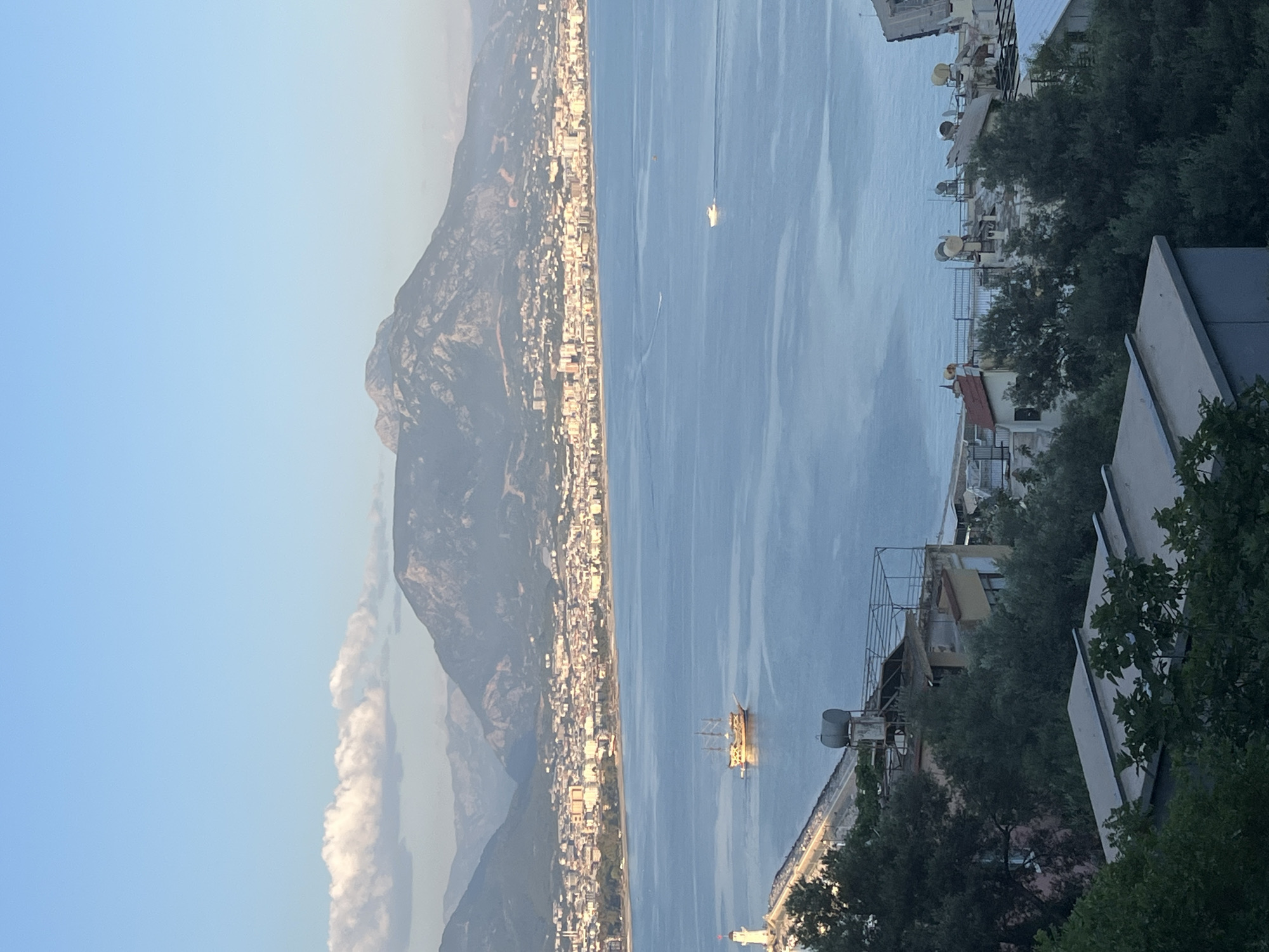 This is a daytime view of the coastline in Alanya, Turkey. A mountain range with cloud cover is to the left. Along the shore is a dense urban area. The calm sea occupies the majority of the scene. Two boats, one with multiple sails, are visible in the water. Buildings are situated on the right.