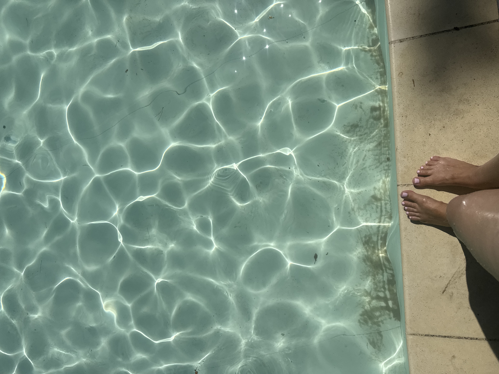 A top-down, close-up shot shows the sparkling surface of a swimming pool. Sunlight refracts through the water, creating a pattern of light and shadow on the pool floor. The water is a clear, turquoise hue, with a slight haze visible in some areas, possibly indicating slight cloudiness or debris. Along the bottom edge of the frame, a beige, textured pool coping is visible. On the coping, the bare feet of a person are positioned, with toes pointing towards the water. The feet appear wet, and the toenails are painted a light pink. A faint shadow is cast on the coping from an unseen object to the left. The scene is devoid of any specific landmarks or architectural features that would indicate a precise location within Orlivshchyna, Ukraine.