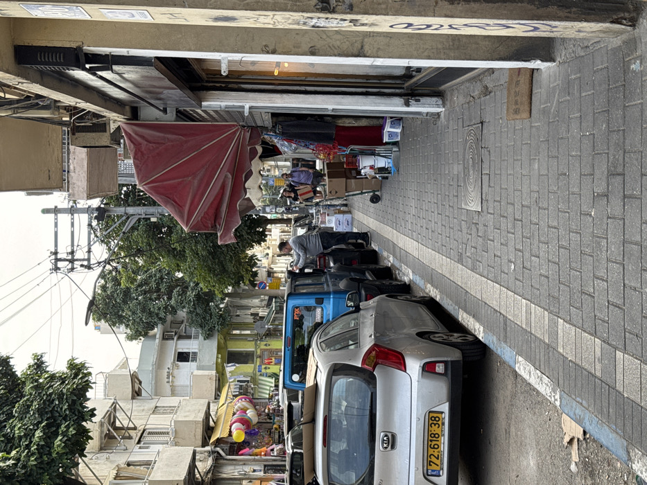 The image depicts an urban street scene in Tel-Aviv, Israel, on an overcast day. A paved sidewalk occupies the right foreground, bordered by a street marked with blue and white curb paint. A silver Kia Picanto, bearing Israeli license plate "72:618:38," is parked closest to the viewer on the street, with cardboard on its roof. Immediately behind it is a blue Jeep Wrangler, with a "FOX" decal on its rear window. A man in a grey long-sleeved shirt is standing beside the driver's side of the Jeep, appearing to attend to something inside or on the vehicle.
To the right, a storefront features a partially folded red fabric awning. Merchandise, including cardboard boxes stacked on a green hand truck, paint buckets, and various items, is displayed on the sidewalk outside the shop. A brown doormat rests near the entrance. Further down the sidewalk, two pedestrians, a woman in a black top and shorts carrying a bag, and a man in a purple shirt with a shoulder bag, are walking away from the camera.
Buildings with multiple stories line both sides of the street, exhibiting a somewhat worn appearance. Overhead utility wires and a street light pole are visible against the cloudy sky. Further down the street on the left, another shop displays colorful balloons and other items outdoors. Graffiti is present on a concrete pillar in the far right foreground, and a circular patterned manhole cover is set into the sidewalk.