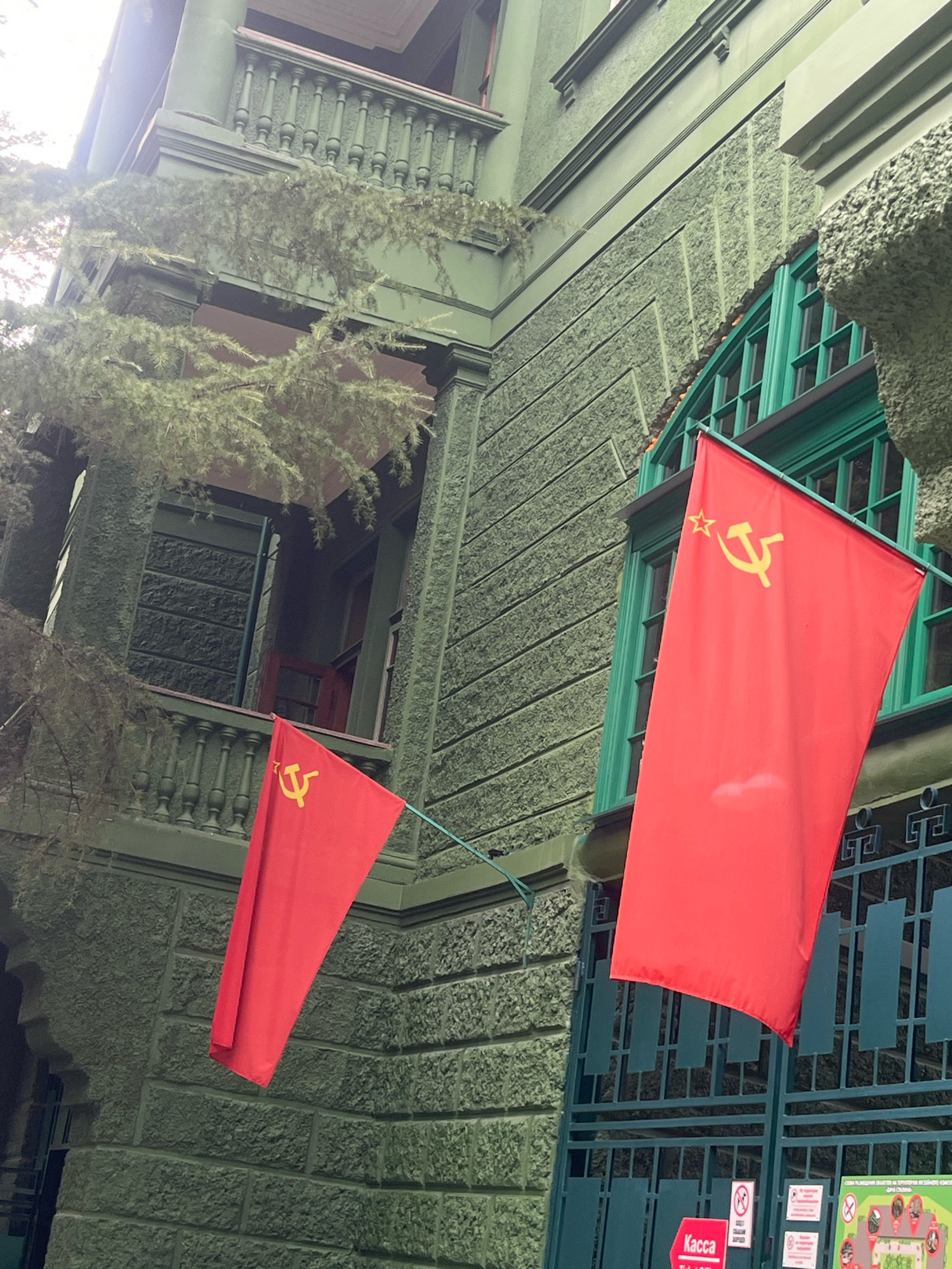 The image shows a two-story, light green building with a textured facade. Two red flags with the Soviet hammer and sickle emblem are hanging from the building. One flag is larger and hangs to the right, while a smaller one hangs to the left. The building has green-trimmed windows, a balcony with decorative railings on the upper floor, and a large green metal gate at the bottom. The setting appears to be outdoors, with some sparse foliage visible in the foreground, suggesting a cloudy or overcast day. The architectural style and the flags suggest a location with historical ties to the Soviet era. This scene is located at the Museum of the History of the Sochi City Resort in Sochi, Russia.