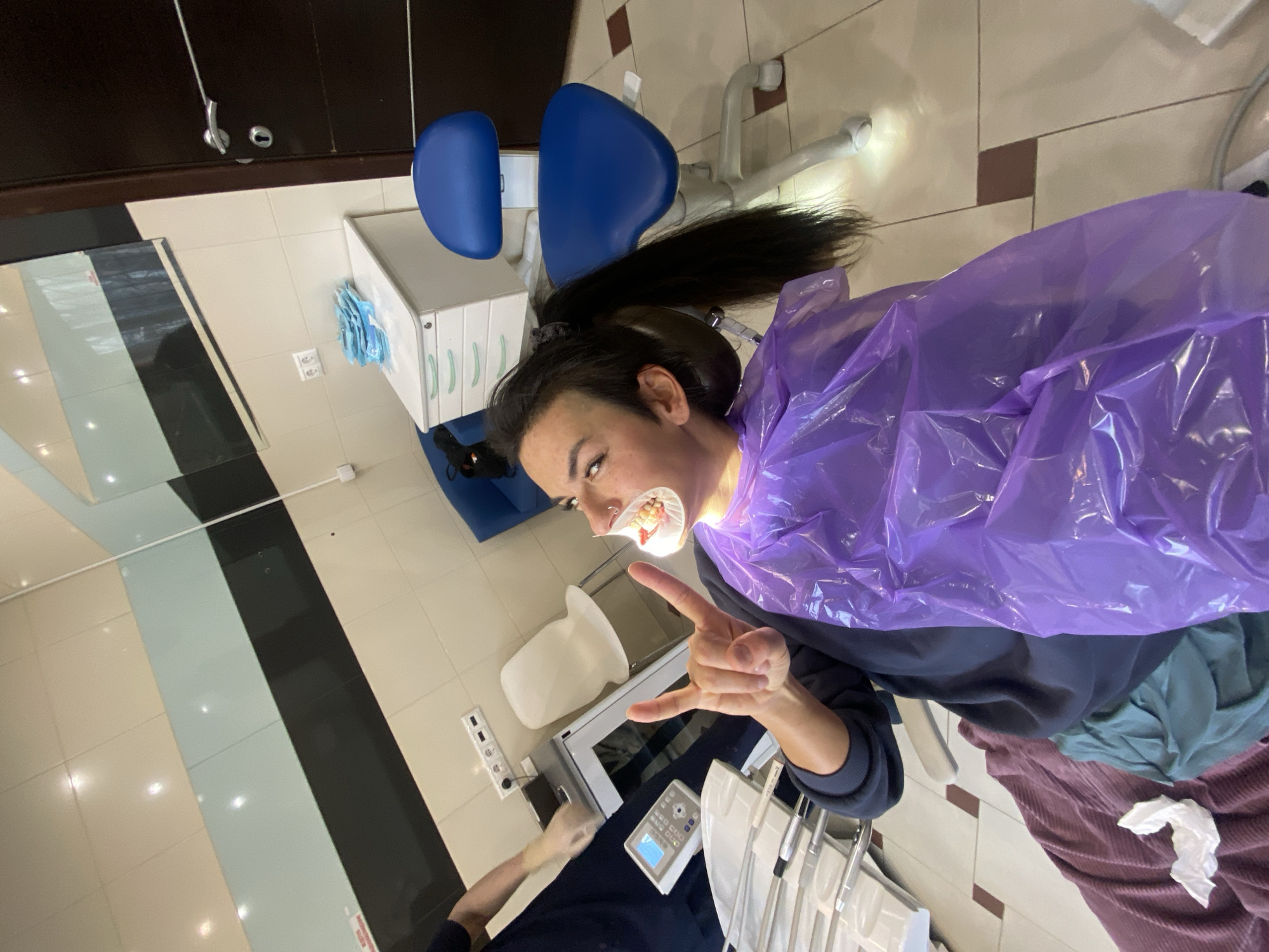 A young woman with a dental retractor in her mouth is lying in a dentist's chair, covered with a purple disposable drape. She is making a "rock on" hand gesture. The setting appears to be a dentist's office, with a dental chair, cabinet, and various dental instruments visible. The location is Moscow, Russia.