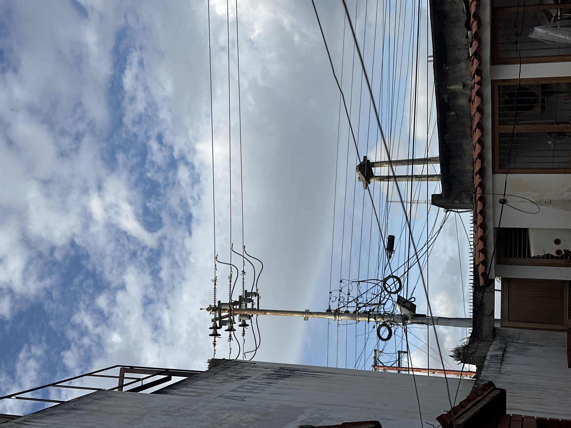This image captures a vibrant, upward-looking perspective of an urban or semi-urban landscape in Juan Griego, Venezuela, dominated by a complex network of overhead utility lines and poles against a partly cloudy sky. The perspective is from below, looking up towards the sky and the structures that punctuate it.
Numerous electrical and communication wires crisscross the frame, creating a dense pattern against the sky. A prominent utility pole stands on the left side, featuring multiple insulators and a concentrated cluster of black wires and equipment. Another pole, further in the background and to the right, also supports a significant amount of cabling.
Parts of buildings or rooftops are visible along the bottom and right edges of the frame. On the right, a building displays distinct terracotta roof tiles, windows with wooden frames and security bars, and an air conditioning unit. Another concrete structure with what appears to be exposed rebar or framework is partially visible in the bottom-left corner. The upper portion of the image is filled with a bright blue sky dotted with scattered white and light gray clouds, indicating daytime. There are no people visible, nor any specific activities taking place. No discernible text is present in the scene, which primarily focuses on the intricate urban infrastructure of power and communication lines.