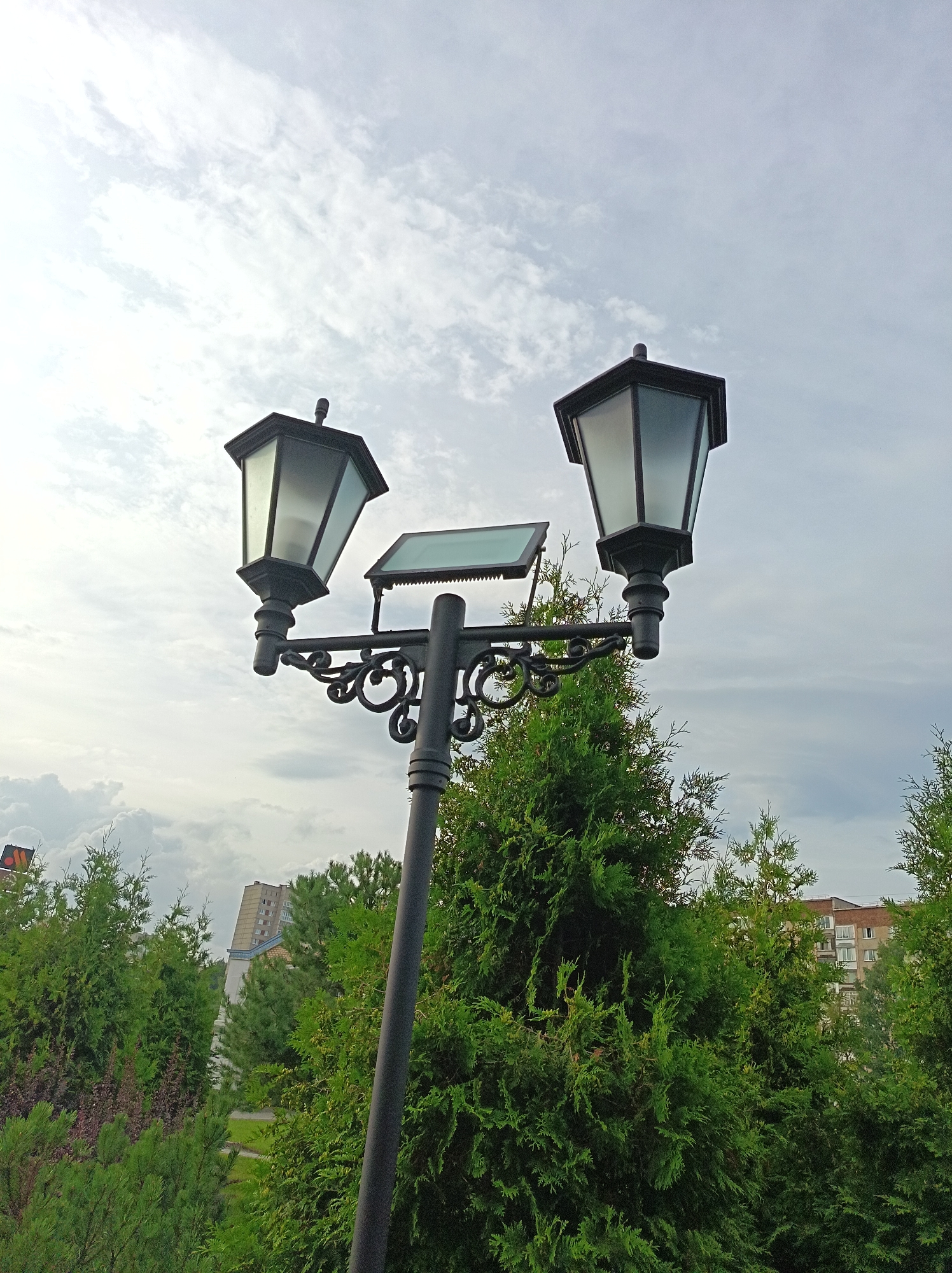 A dual-headed lamppost with a solar panel is pictured against a cloudy sky. The lamppost is black and has decorative scrollwork. Each lamp is hexagonal with frosted glass panels and a black cap. In the background, a building with many windows is visible, surrounded by lush green trees. The scene appears to be in an urban park or residential area.