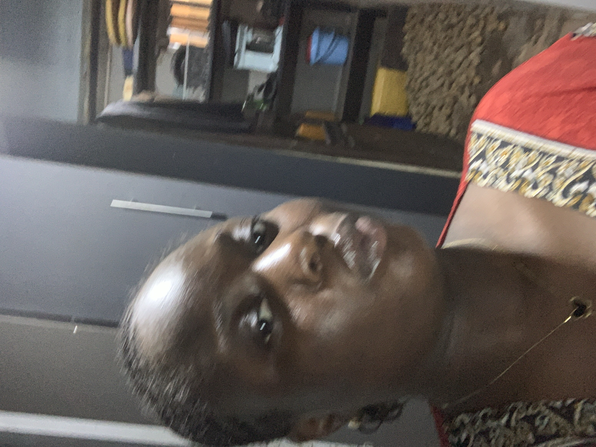 A woman is taking a selfie indoors. She has dark skin and dark hair. She is wearing a red dress with a patterned neckline. She has a necklace with a clover-shaped pendant. The background appears to be a room with shelves filled with various items. The lighting suggests it is daytime. The location is not specified.