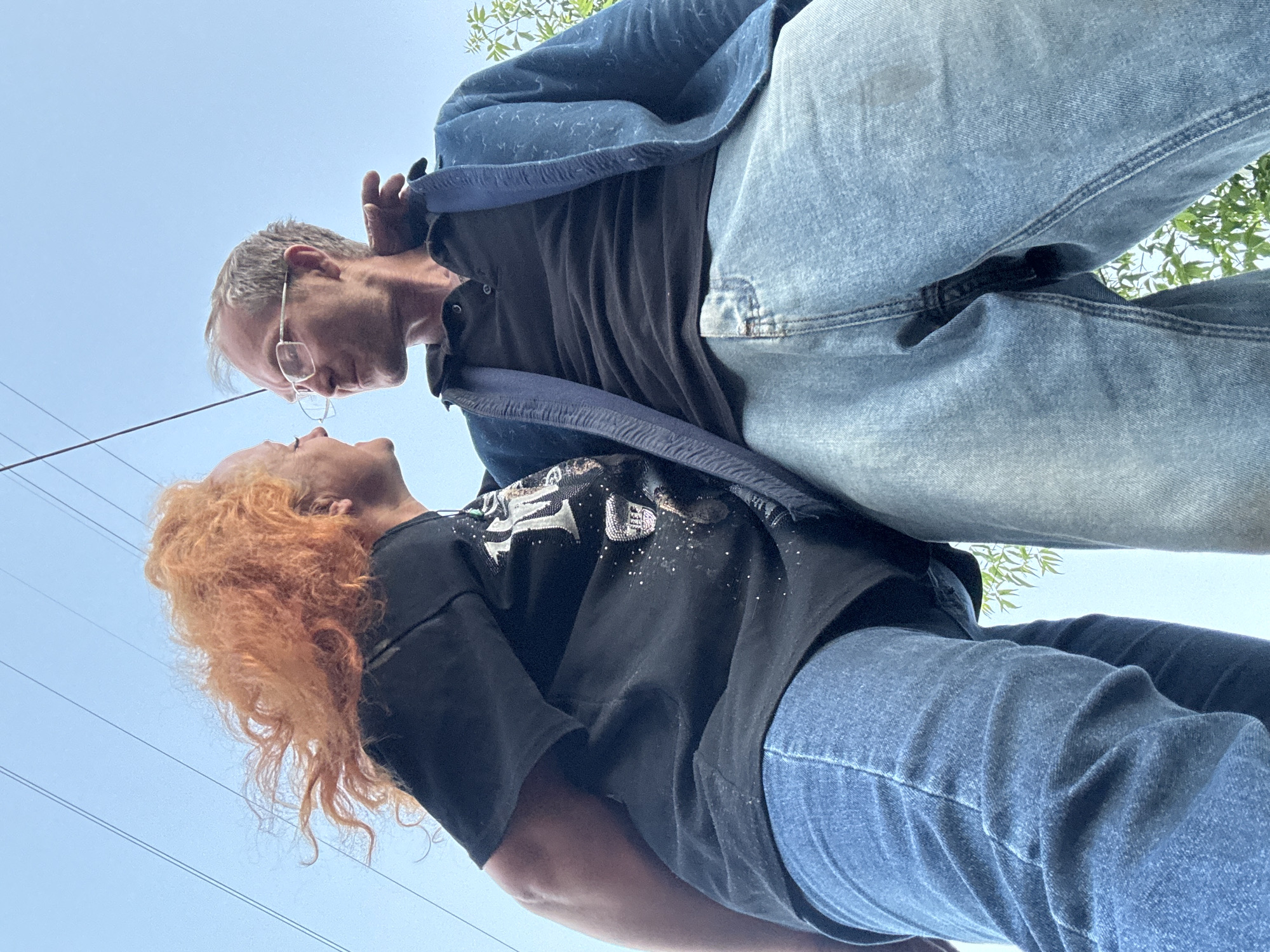 Captured from a low-angle perspective, two individuals are seen from below, framed against a clear, bright blue sky with sparse green foliage visible in the upper and lower right corners. The composition emphasizes their closeness and an intimate moment.
The subject on the left is a man with grey, receding hair and glasses, wearing a dark t-shirt, a blue denim jacket, and light blue jeans. He is looking down and leaning towards the other person. The subject on the right is a woman with long, curly, reddish-orange hair, wearing a black t-shirt with a sparkly white graphic, and dark blue jeans. She is looking upwards, meeting the man's gaze. Their faces are very close, almost touching, suggesting an affectionate interaction, possibly leading to a kiss or a whispered conversation.
Thin, dark power lines are visible stretching across the blue sky, particularly near the woman's head. The woman's black t-shirt features a graphic design with the partial text "EAGLES" visible, adorned with glittery or metallic accents.
The scene takes place outdoors on a clear, sunny day, likely midday or afternoon, indicated by the bright, even lighting and cloudless sky. The overall impression is one of connection and tenderness between the two subjects in an open-air setting in Chaikovsky, Russia.