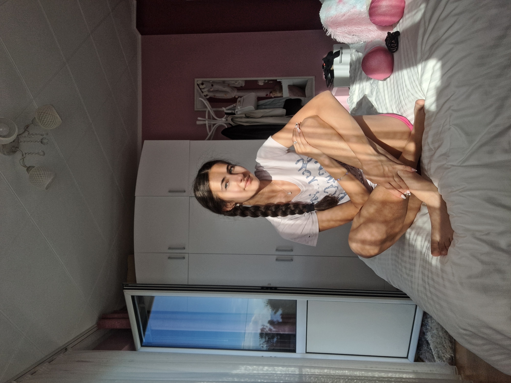 A young woman is sitting on a white bed. The room is lit by sunlight shining through the window. The room includes a closet, various accessories, and toys on the bed. She is wearing a white T-shirt and has two braids.