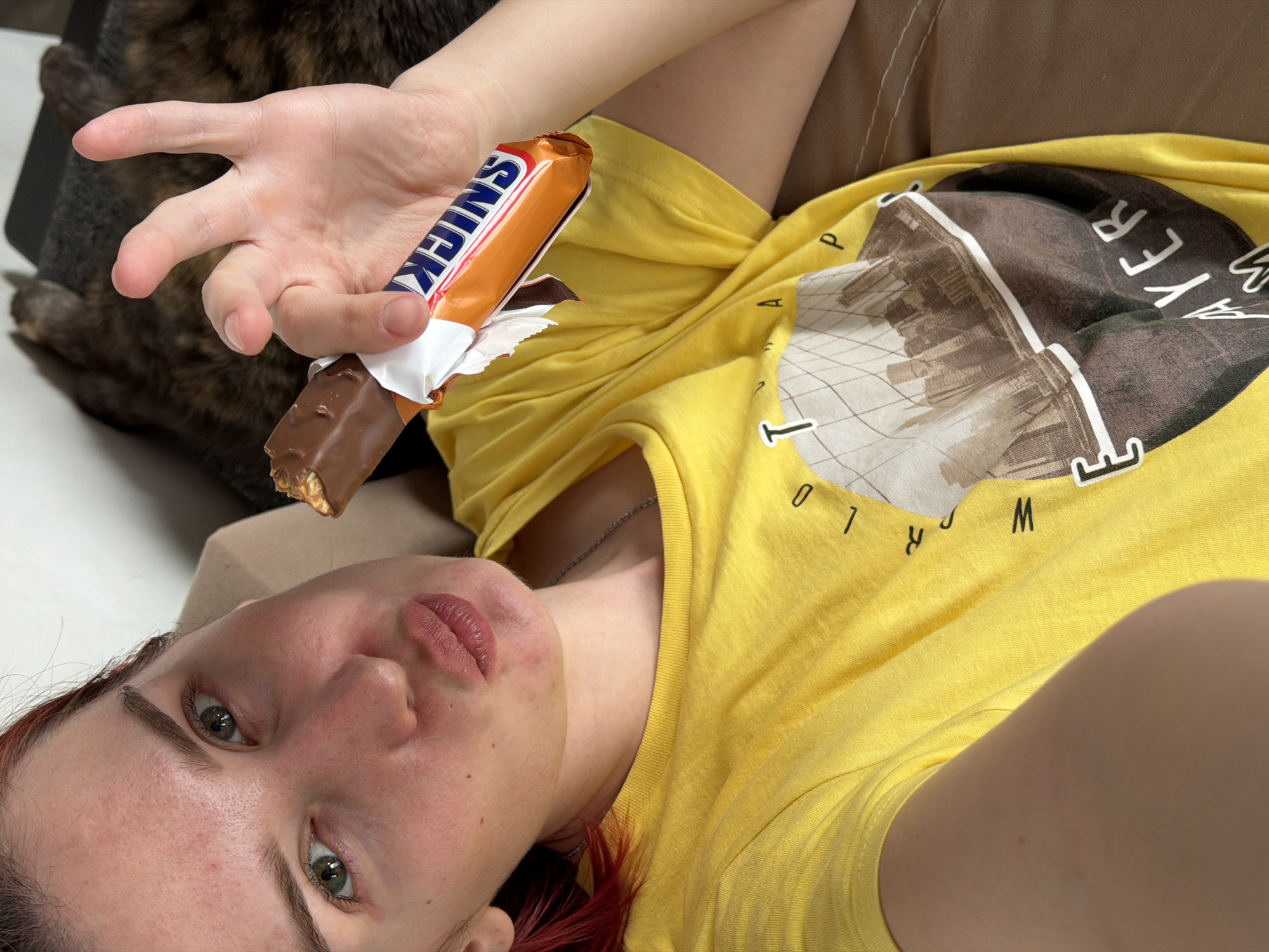 A close-up, eye-level shot captures a woman lying down indoors, looking towards the camera. She has fair skin, green eyes, and reddish hair, with her lips slightly parted in a neutral, relaxed expression. She is wearing a bright yellow t-shirt featuring a brown and white cityscape graphic. Fragmented black text is also visible on the shirt, including letters such as "W", "O", "R", "L", "D", "Y", and "E", "R". A delicate silver chain necklace adorns her neck.
Her left hand is raised, holding a partially eaten Snickers chocolate bar, with the brand name "SNICKERS" clearly visible in white and blue lettering on the orange wrapper. In the blurry background to the left of her hand, a dark-furred animal, possibly a cat or dog, is partially visible. The overall setting appears to be a casual indoor environment, likely a bed or sofa, suggested by the soft, textured brown and white fabric in the background. The lighting indicates it is daytime. The image conveys a relaxed and comfortable moment in Chaikovsky, Russia.
