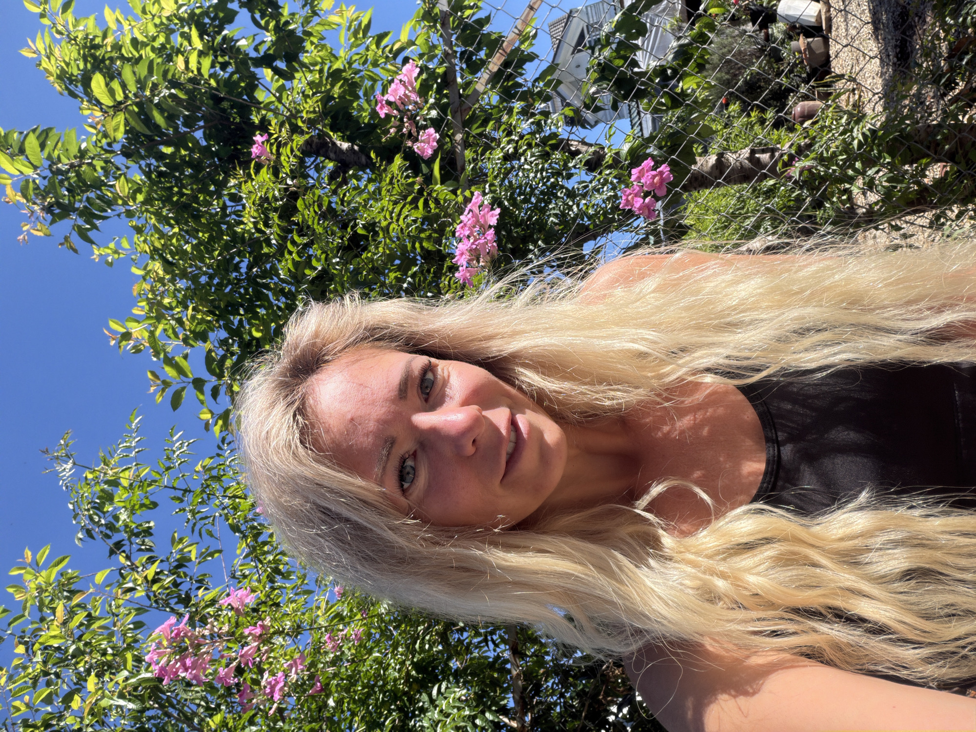 A Caucasian woman with long blonde hair and blue eyes is taking a selfie outdoors. She is wearing a black tank top. Behind her, a tree with green leaves and pink flowers is visible, along with a chain-link fence and a clear blue sky. The image appears to be taken in a residential area, with some buildings and potted plants in the background. The lighting suggests it is daytime, likely sunny. The location is the city of Da Lat, Vietnam.