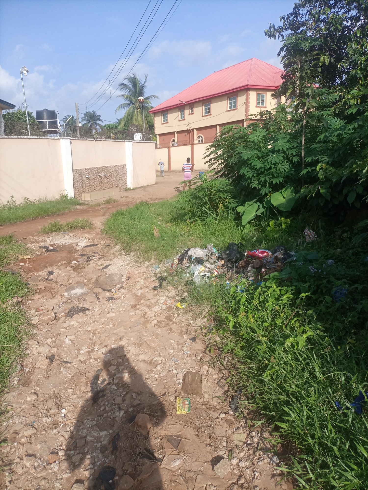 The image shows a dirt path in Nkpor, Nigeria, with a pile of garbage on the right side of the path. In the background, there is a two-story building with a red roof and a beige exterior. A person wearing a striped shirt and pink shorts is walking away from the camera. The sky is blue with some clouds. The scene appears to be in a residential area, with vegetation and a fence visible. A shadow of the photographer is cast on the dirt path in the foreground.