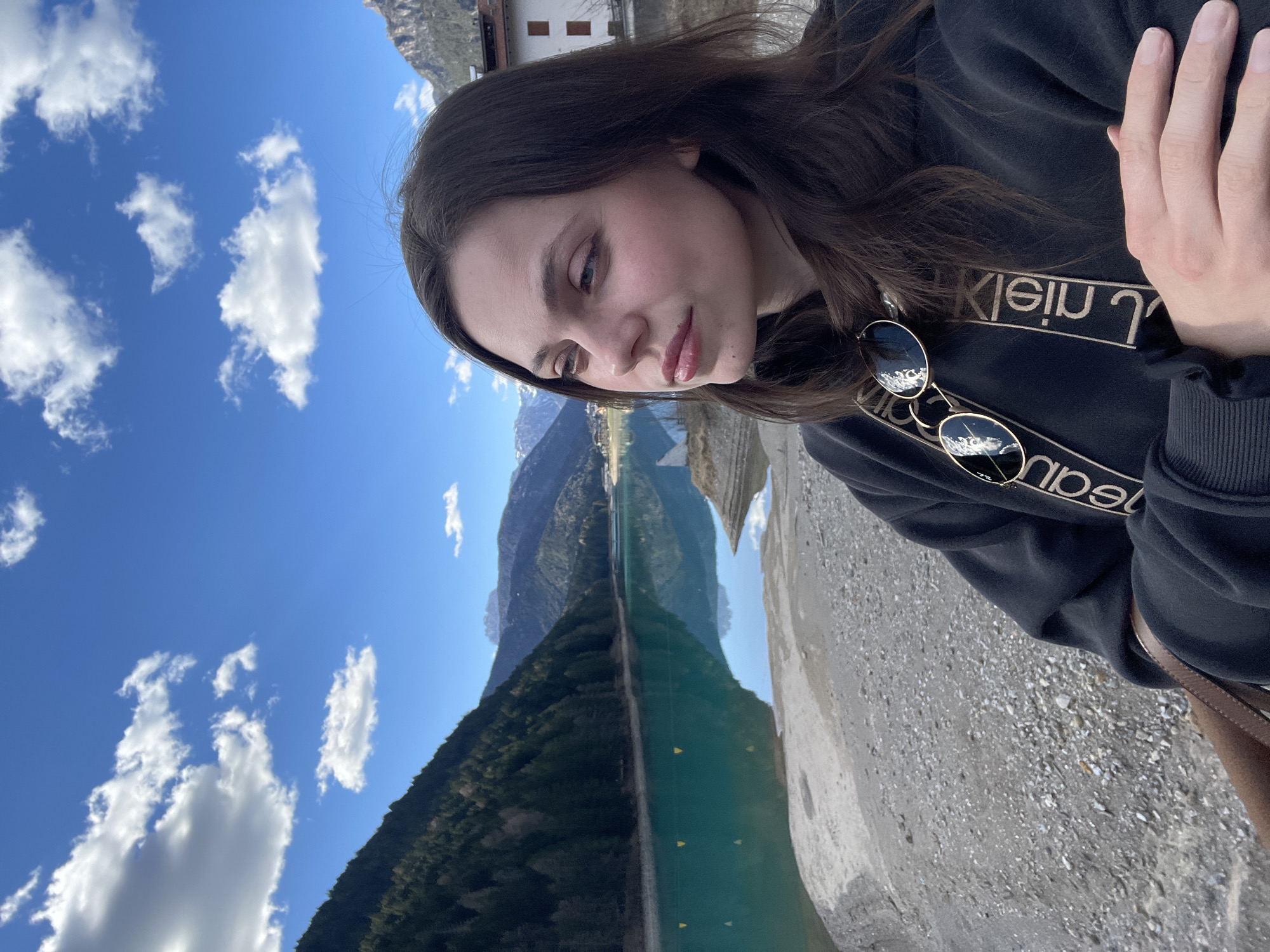 A woman is the focus of a selfie-style photograph. She has long brown hair, and is wearing a black Calvin Klein sweatshirt and sunglasses. Her hands are crossed in front of her. The background shows a landscape with a lake, mountains, blue sky, and scattered clouds. The location appears to be in Auronzo di Cadore, Italy, based on the mountainous landscape. The photo is oriented sideways. A building is visible in the background.