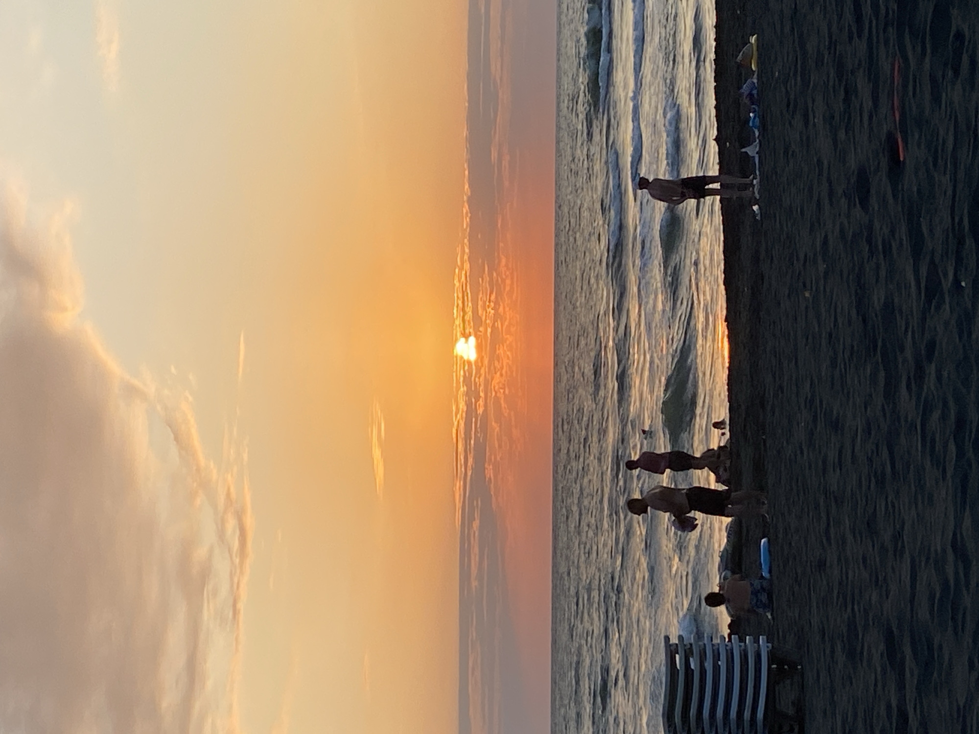 This image depicts a sunset scene on the beach in Ureki, Georgia. The sky is filled with warm orange and pink hues, with clouds tinged by the setting sun. The sun itself is partially obscured by a horizontal band of clouds, creating a soft glow over the horizon. The sea in the foreground is moderately active with waves breaking near the shore.
On the dark sand beach, several individuals are visible. To the left, a stack of white lounge chairs is neatly arranged. Closer to the water, a person is seated on the sand. Further to the right, a man is standing, holding what appears to be a light-colored object, possibly a towel or clothing item. Another person is visible in the water further out, and another individual stands on the beach with their back to the camera. Scattered items like towels and bags are visible on the sand near some of the people. The overall atmosphere is that of a relaxed evening on a beach.