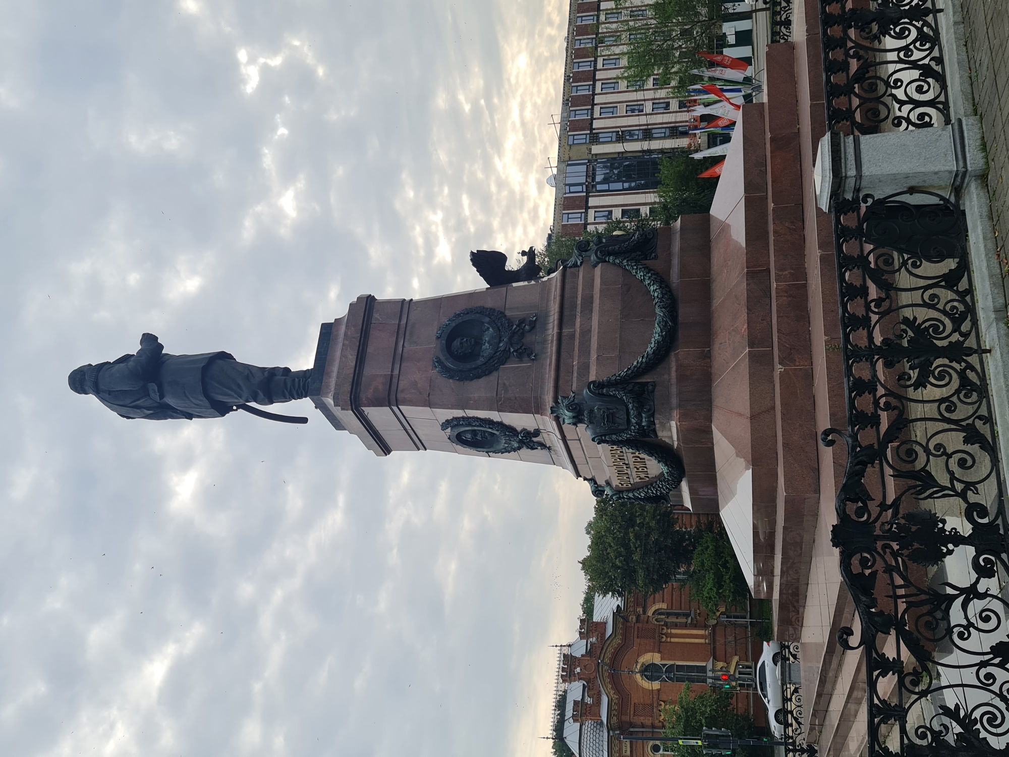 A low-angle, full shot shows a large monument to Grigory Shelikhov in the city of Irkutsk, Russia, on a cloudy day. The monument, made of dark bronze and pink granite, stands tall with a statue of a man in uniform at its apex, holding a sword. The monument's base is adorned with bas-reliefs of wreaths, portraits, and inscriptions. To the left of the monument is a red brick building with ornate architecture. To the right, a modern building with large windows is under construction. In the foreground, a black wrought-iron fence with ornate scrollwork separates the monument from a paved walkway. A white car is visible on a street to the left, and several flags are planted in the ground behind the monument. Birds can be seen flying in the sky.