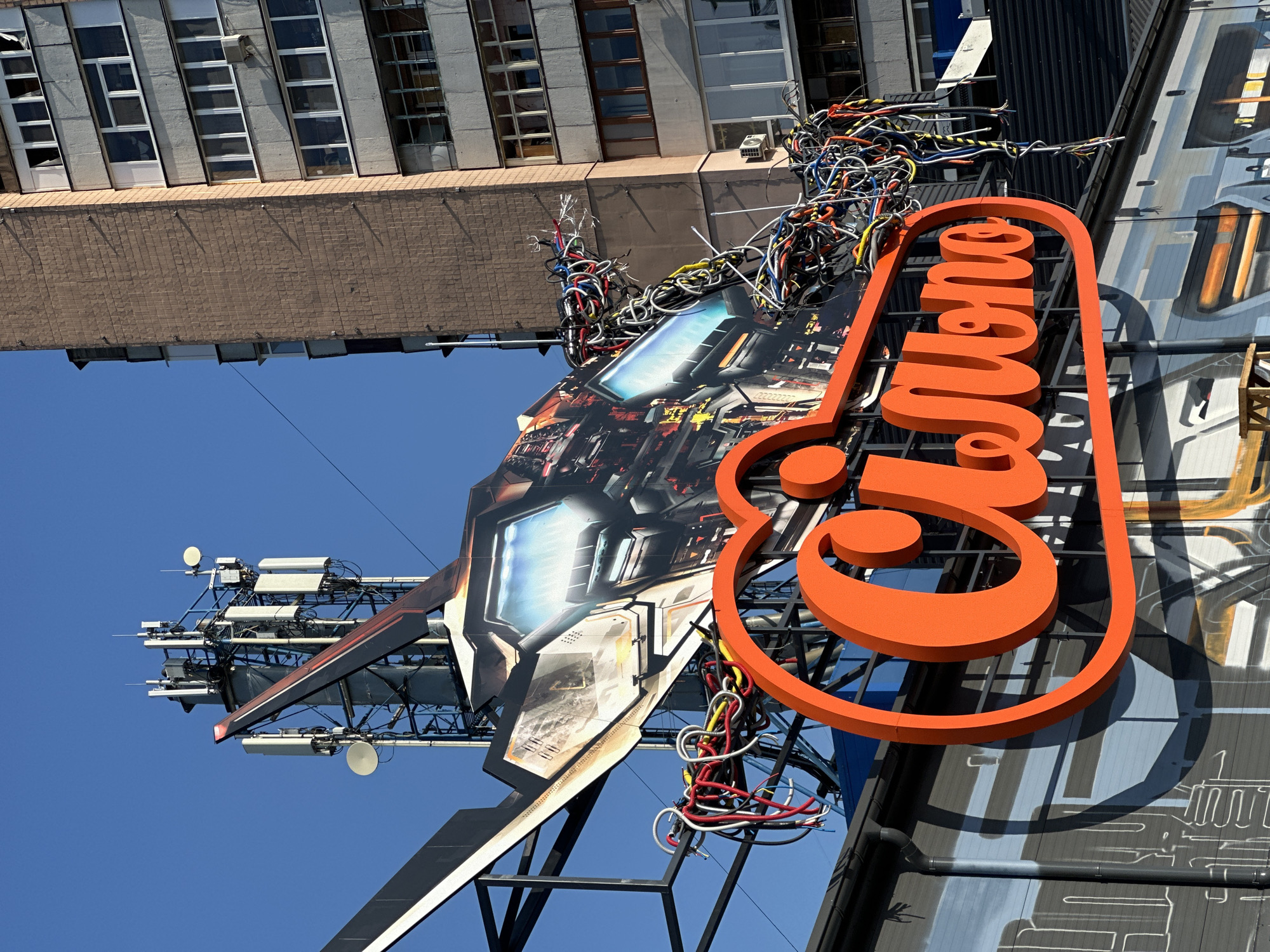 A close-up, eye-level outdoor shot shows a vibrant orange sign with large, stylized lettering that reads "Cilmon" against a backdrop of a building and a cell tower. The sign is positioned on the left side of the frame, and the building, a multi-story residential structure with a brick facade and numerous windows, is on the right. The cell tower, a tall, industrial metal structure with multiple antennas and dishes, is visible behind and to the left of the building. The foreground of the image is dominated by the bright orange sign, which has a sleek, modern design. Behind and above the sign, a portion of a large, futuristic-looking graphic, possibly part of a billboard or mural, is visible. This graphic appears to depict a metallic object with glowing blue elements and is adorned with a tangle of colorful electrical wires. The sky above is a clear, deep blue, suggesting a sunny day. The overall impression is one of urban dynamism, with a blend of old and new architecture, technology, and bold signage.