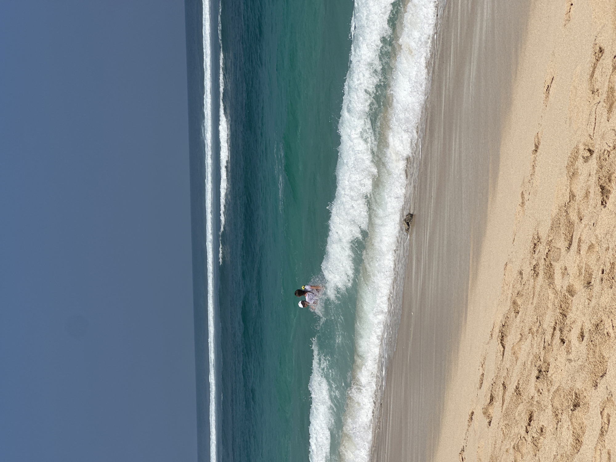 The image depicts a beach scene in Kuta Selatan, Indonesia. In the turquoise water, two individuals are visible, appearing to be playing or swimming amidst crashing waves. One individual, wearing a white shirt and a white hat, is partially obscured by the water. The other individual, with dark hair and wearing a white shirt, is facing away from the camera. The foreground shows a sandy beach with footprints, and the background features the vast ocean with multiple lines of waves and a clear blue sky.