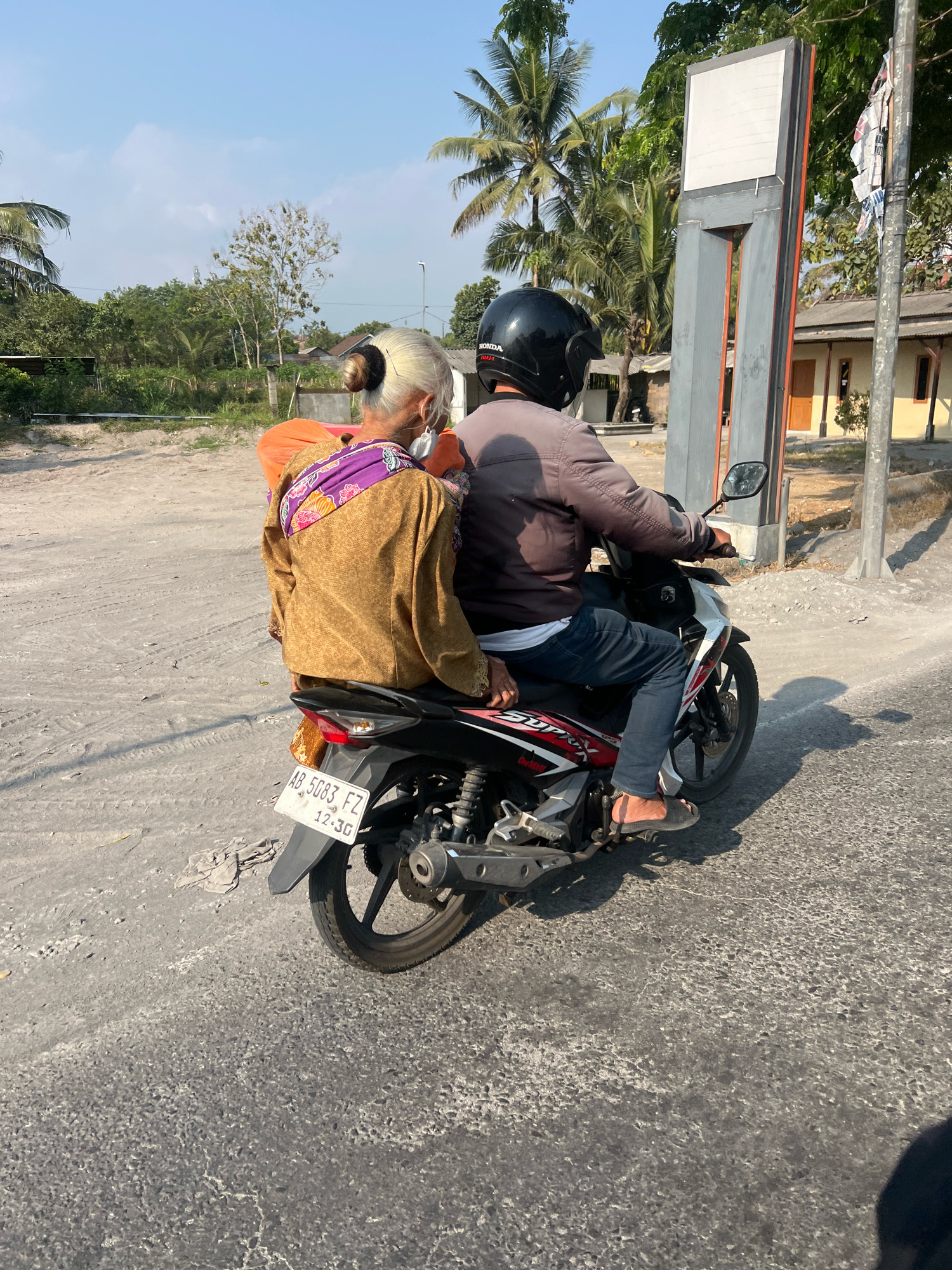 Two people are riding a motorcycle on a dirt road in Tempel, Indonesia. The person in the back is an elderly woman with her hair in a bun, wearing a brown jacket and a purple sash. She is holding an orange bag. The person in the front is a man wearing a black helmet and a gray jacket. The motorcycle has a license plate that reads "AB 5083 FZ". In the background, there are palm trees, a building, and a sign. The sky is clear and blue.