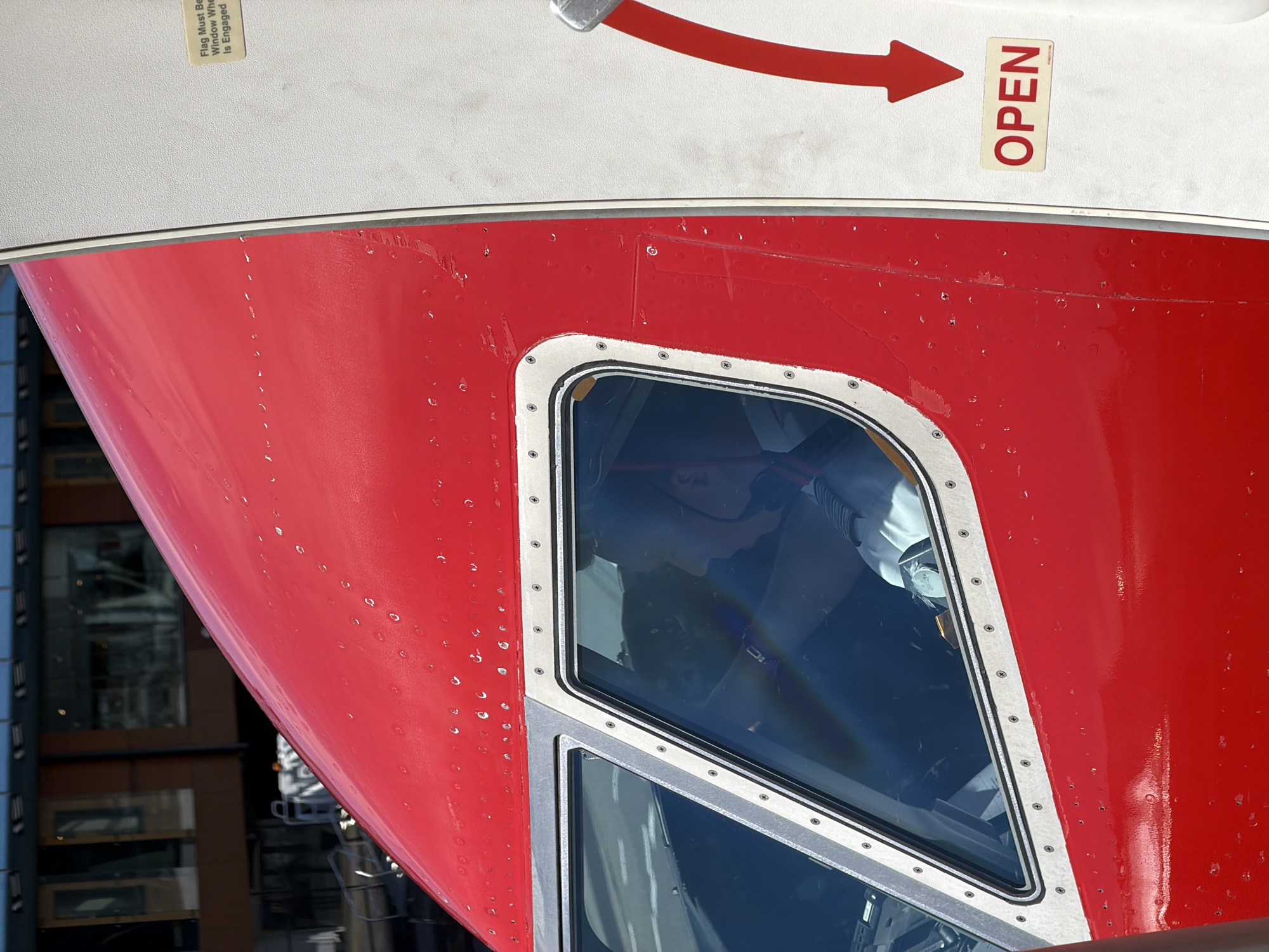 A close-up shot of the cockpit of a red aircraft shows a pilot wearing a headset, looking out the window. The pilot is seated and positioned behind the control yoke, with their profile visible through the reinforced glass. The aircraft's fuselage, painted a vibrant red, dominates the foreground, with visible rivets and some minor scuff marks. To the right of the fuselage, a white door is partially visible, marked with a red downward-pointing arrow and an "OPEN" sign. The background reveals a blurred view of buildings with large windows, suggesting an airport terminal environment. The overall lighting indicates it is daytime. The image does not provide specific clues to pinpoint the location within Tallinn, Estonia, beyond it being at an airport.