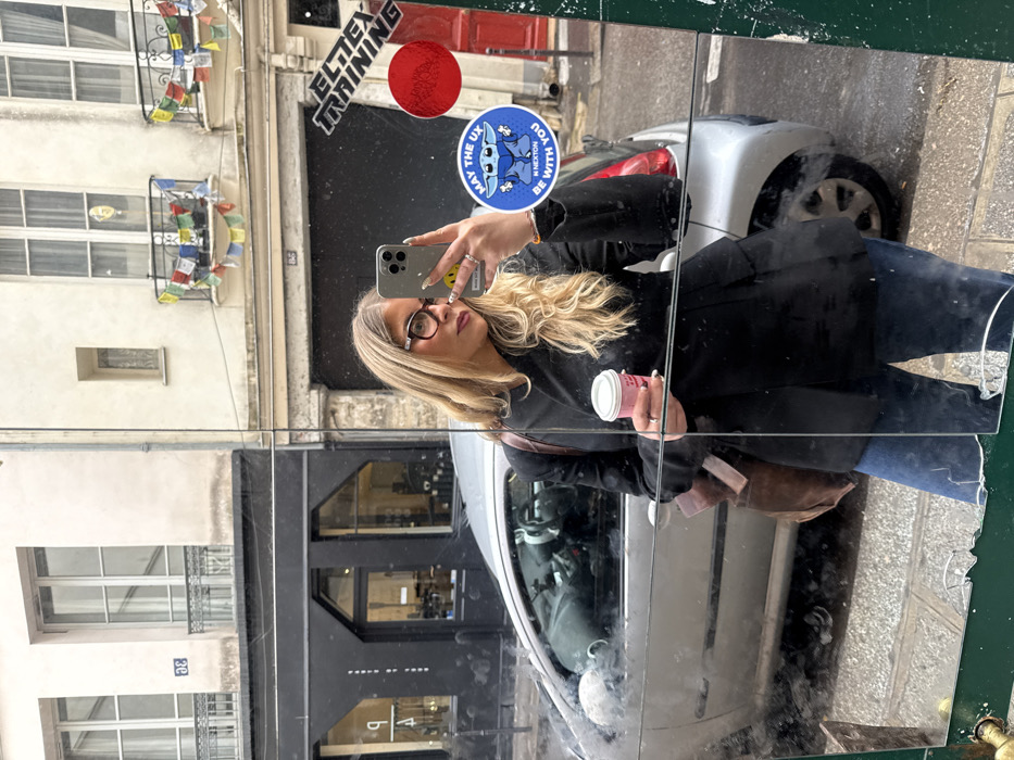 A blonde female, estimated to be in her 20s or 30s, captures a mirror selfie on a street in Paris, France. She wears dark-rimmed glasses, a black blazer, and blue jeans, holding a light pink paper coffee cup in her left hand and an iPhone 14 Pro/Pro Max in her right hand to record her reflection. A ring is visible on her left ring finger and her right index finger. A brown bag strap rests on her left shoulder.
The mirror, appearing weathered with cracks in the lower right corner and streaks of grime across its surface, reflects a typical Parisian streetscape. To the left, a light-colored building bears the number "33" above a dark-fronted shop. To the right, a similar light-colored building, numbered "35," features a balcony decorated with colorful Tibetan prayer flags, positioned above another dark shopfront. A silver two-door hatchback car, consistent with a Peugeot 206, is parked on the street behind the woman. Two stickers are affixed to the mirror's surface: a white text "ELTEX TRAINING" sticker positioned above a plain red circular sticker, and a blue circular sticker featuring a cartoon Grogu and the text "MAY THE UX BE WITH YOU". The street surface is paved, and the overall lighting suggests an overcast day.