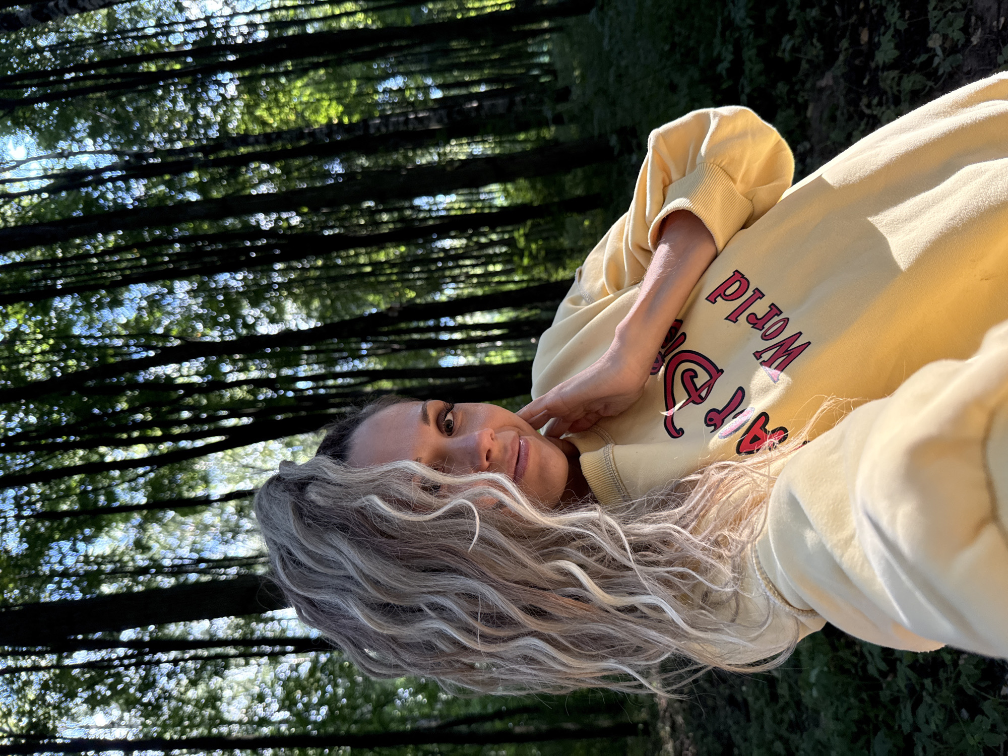 A woman with blonde, wavy hair pulled into a high ponytail is captured in a selfie taken outdoors. She is wearing a yellow sweatshirt with red lettering that says "Disney World". Her left hand is resting on her jawline. The background consists of a dense forest with tall trees and dappled sunlight filtering through the leaves. The image appears to have been taken during the daytime. The location is identified as Dumino, Russia.