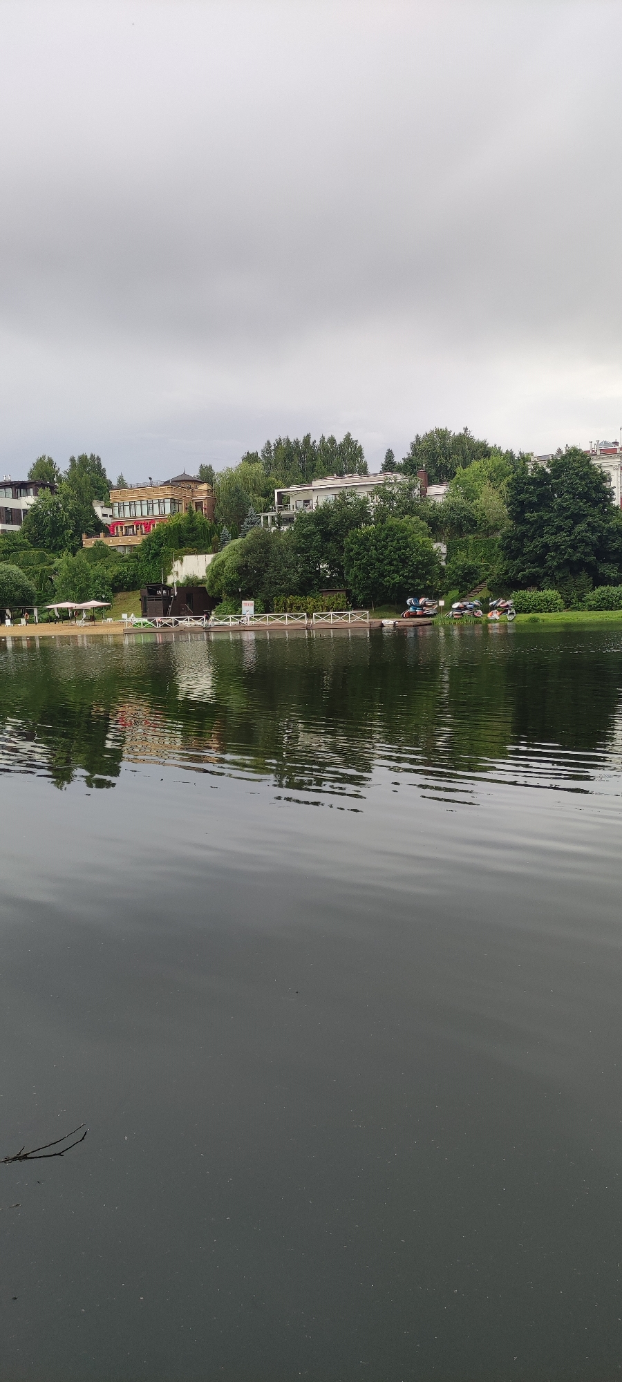 The image depicts a cloudy day scene at a waterfront in Angelovo, Russia. In the foreground, the dark water of a lake or river fills the lower half of the frame, with subtle ripples distorting the reflection of the shore. A small, thin branch floats on the surface in the lower left corner. The middle ground reveals a sandy beach area with several white beach umbrellas and lounge chairs. Adjacent to the beach is a wooden dock with white railings. On the dock and further along the shore, several small boats, some with people near them, are visible, as well as some wheeled carts. The background is dominated by lush green trees and several multi-story buildings, characteristic of a residential or resort area. The buildings have a modern architectural style with various colors and materials. The sky is overcast and light gray, indicating a subdued lighting condition.