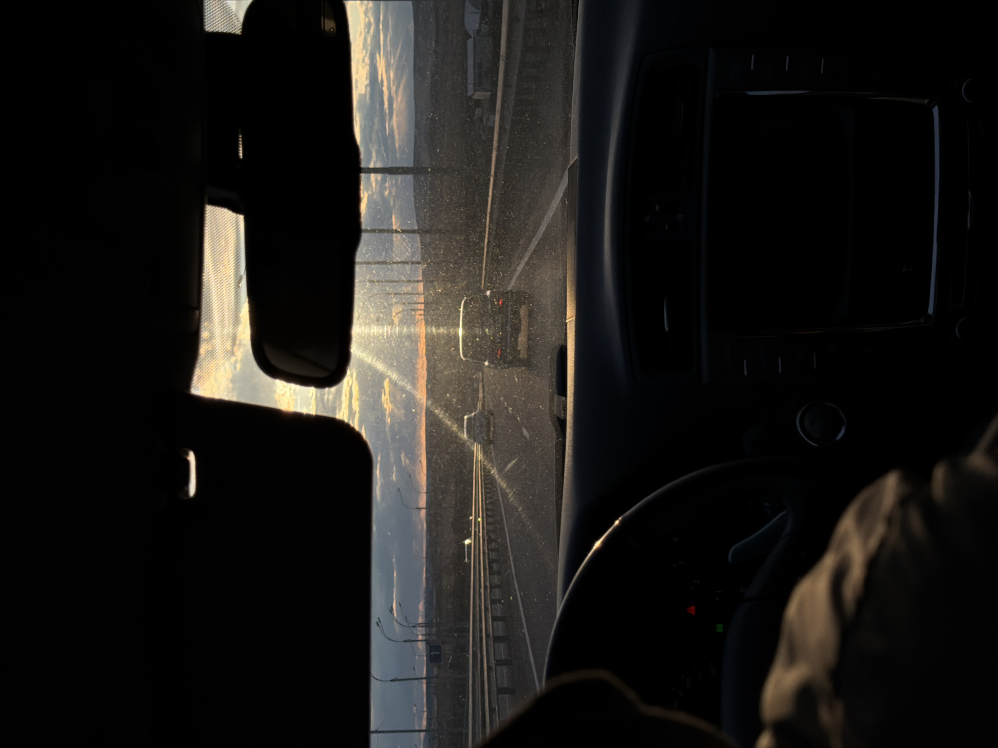 From inside a car, a view is seen looking forward. The sky is bright from the sun, and the road ahead is a highway with a car in the distance. To the left is a rearview mirror. To the right is a dashboard, steering wheel, and a person’s arm. It is not possible to determine from the provided information whether the location is in Krasnoyarsk, Russia.