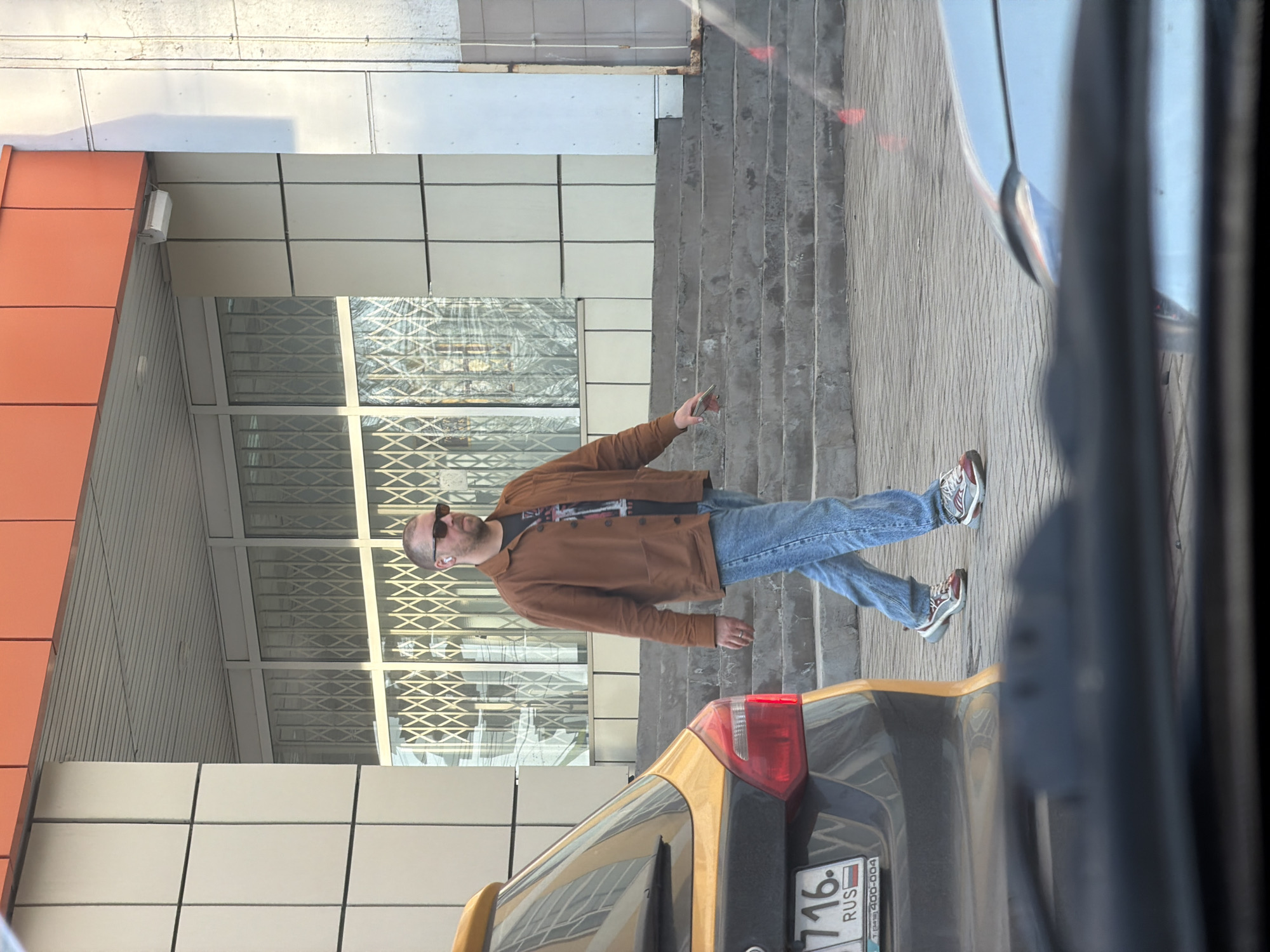 A man stands on a sidewalk in Izhevsk, Russia. He has a shaved head, sunglasses, and an earbud in his left ear. He wears a brown jacket, a dark t-shirt, blue jeans, and sneakers. He holds a phone in his right hand. He is standing in front of a building with large windows and a set of stairs. A car with a yellow taxi sign on the roof is parked nearby. The car has a Russian license plate with the number 716. Another car is partially visible, obscuring the view.