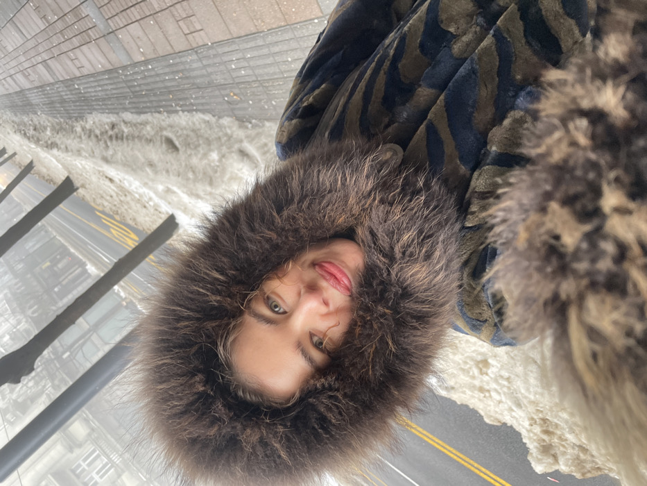 The image depicts a female individual taking a selfie outdoors on a winter day in Belgrade, Serbia. The subject, positioned centrally and angled, wears a dark brown fur-lined hood framing her face, light red lipstick, and a coat featuring a dark blue/black and olive/brown abstract pattern with additional fur trim. The background reveals an urban street environment under hazy conditions. A road with double yellow lines is visible on the left, bordered by a substantial accumulation of snow. To the right, a grey paved sidewalk is partially covered in snow. Bare deciduous trees line the street, and multi-story buildings with classical architecture are faintly discernible through the fog in the distant background. The scene indicates cold, overcast weather with reduced visibility, typical of a winter day in central Belgrade.