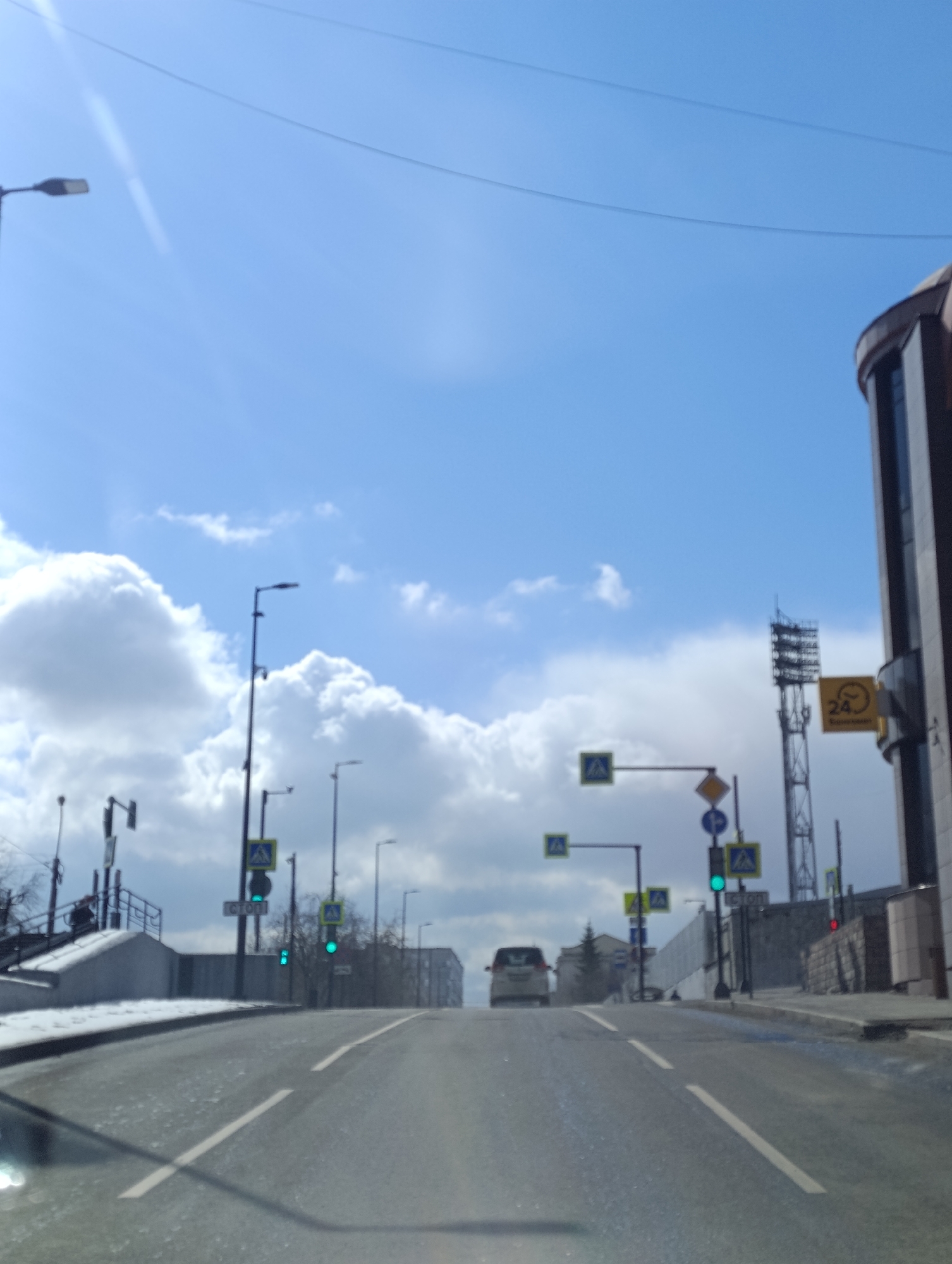 This image captures an ascending road in an urban setting, likely in Krasnoyarsk, Russia, under a bright, partially cloudy sky. The perspective appears to be from inside a vehicle, with the lower part of the windshield or dashboard visible at the bottom.
The asphalt road features white lane markings, including a solid line on the left and a dashed line on the right, indicating traffic flow uphill. Numerous streetlights and utility poles line both sides of the road. Prominent pedestrian crossing signs (blue squares with white triangles depicting a walking figure) are visible, along with other traffic signs such as a yellow diamond-shaped sign and a round blue sign. Several traffic lights are present, with some showing green, and one further ahead on the right appears red. A light-colored car, possibly a white SUV or minivan, is seen driving up the road in the distance.
On the left side of the road, a concrete barrier is partially covered with snow, suggesting cold weather or recent snowfall. On the right, a large, dark-colored building with a curved glass facade stands prominently. Attached to this building is a yellow square sign displaying a black number "24" with an arrow curving around it, indicating a 24-hour service, with faint Cyrillic text below. Behind this building, a tall metal tower with lights, resembling a stadium light tower or communications mast, is visible against the sky. Overhead power lines traverse the upper portion of the frame.
The sky is a bright blue with large, white, cumulus clouds. A lens flare is noticeable in the top-left corner, indicating direct sunlight. The scene depicts a typical street environment with ongoing traffic on a clear, cool day. No people are clearly visible. A white rectangular sign with dark text, possibly "СТОП" (STOP), is partially visible on a pole on the left.