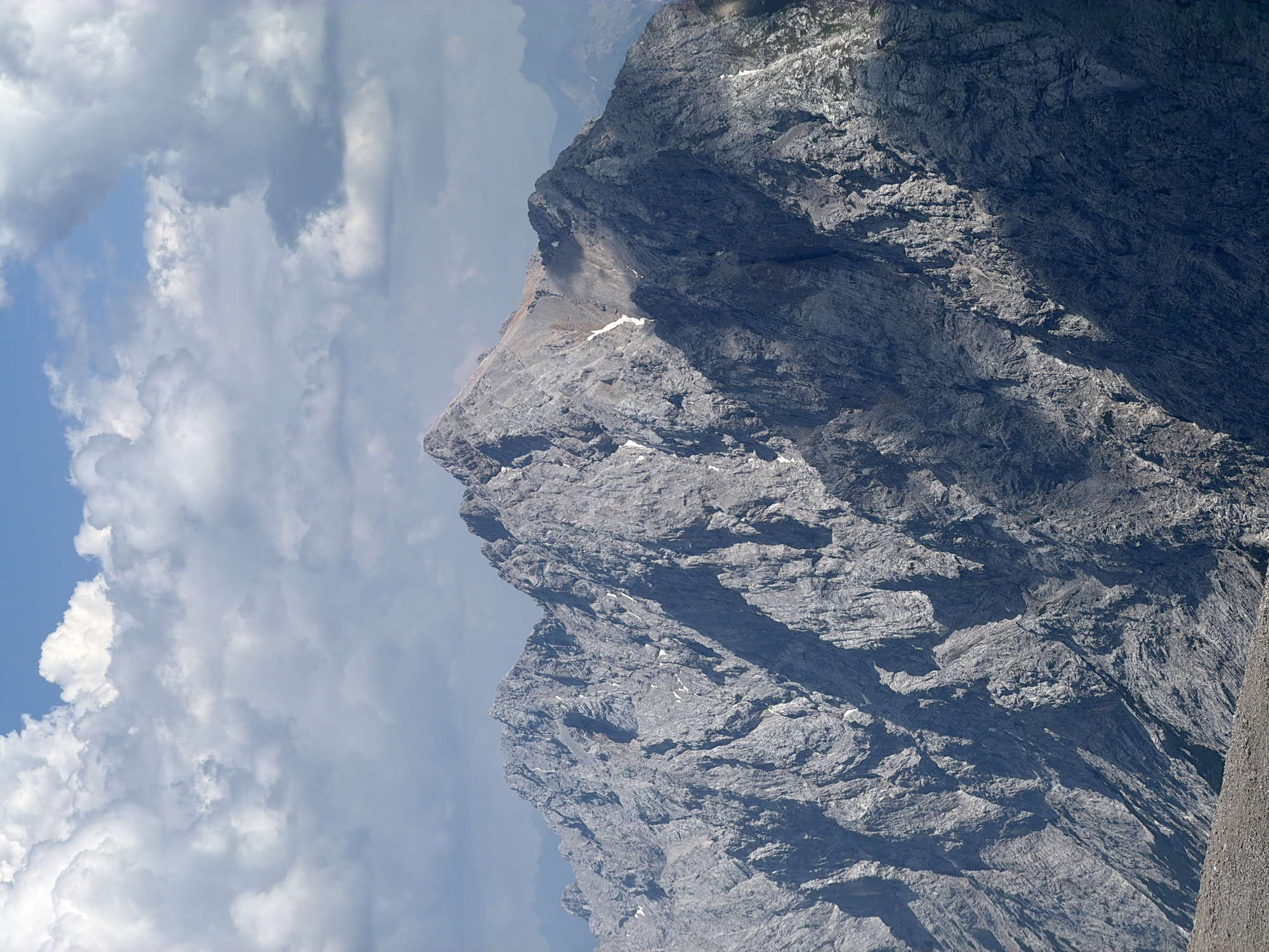 This media file depicts a majestic, rugged mountain landscape under a bright, partly cloudy sky. The primary subject is a sheer, grey rock face, characterized by its intricate texture and dramatic shadows. The mountain appears to be a significant natural landmark, possibly part of the Alps given the location context of Garmisch-Partenkirchen, Germany.
The scene is dominated by the imposing scale of the mountain, which occupies a large portion of the frame. The rock formations are sharp and angular, with visible layers and crevices that speak to the geological processes that shaped them. Patches of white, possibly snow or light-colored rock, are scattered across the mountainside, adding visual interest.
The sky is a dynamic mix of clear blue and fluffy white clouds. The clouds are well-defined, with some casting shadows on the mountain, indicating a time of day when the sun is high. The lighting suggests it is daytime, likely late morning or early afternoon, with the sun casting strong highlights and deep shadows on the mountain's surface. The overall atmosphere is one of natural grandeur and isolation. There is no text visible in the image.