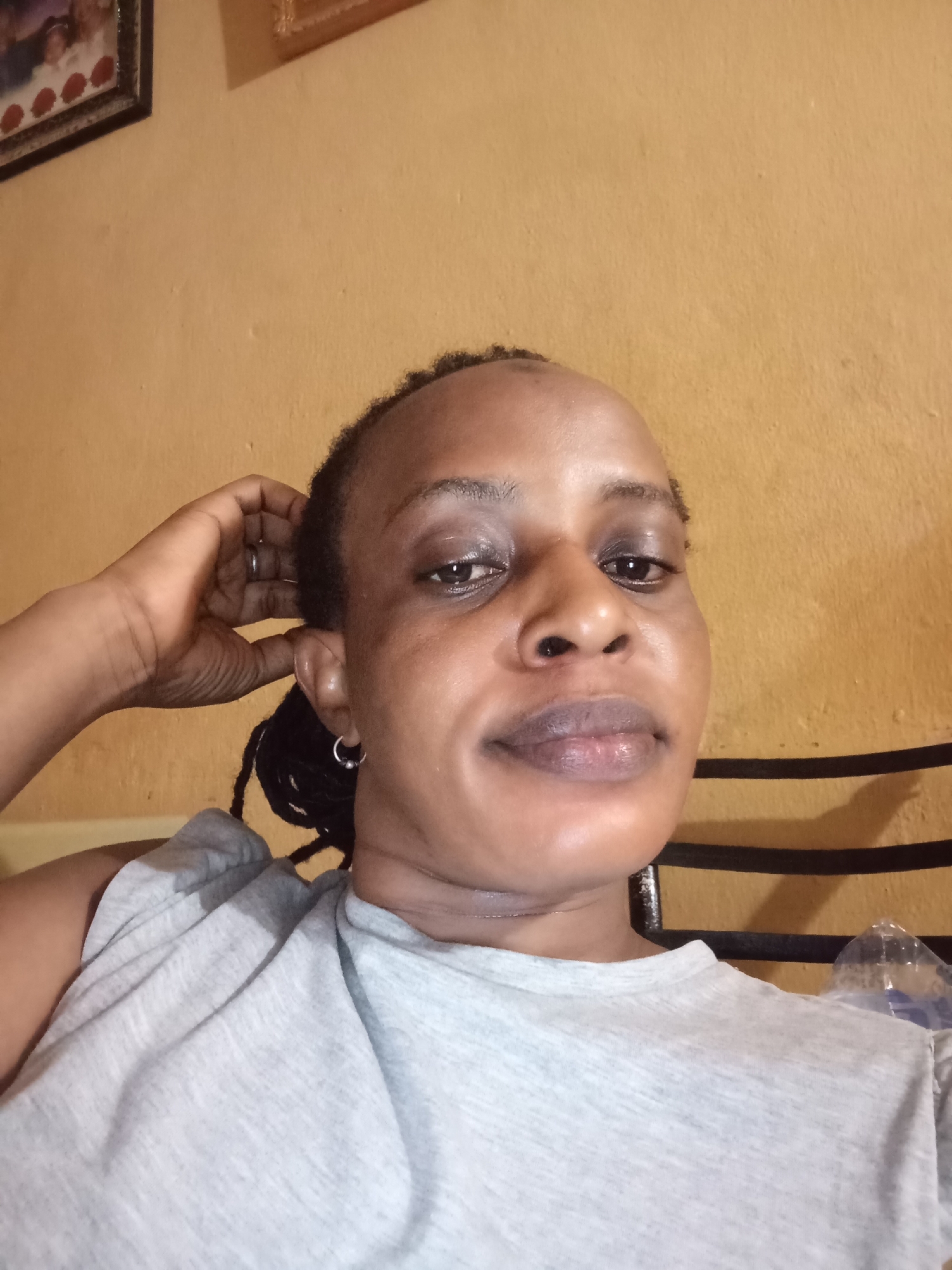 A woman with dark skin and dark hair in braids sits against a light yellow wall. She wears a gray t-shirt. Her left hand is raised to the back of her head. She has a small silver earring in her left ear and a ring on her left hand. Behind her is a metal headboard. A picture frame is visible in the upper left corner of the image. A partially visible wall-mounted decorative object is in the upper center of the image. A discarded plastic water bottle is visible behind the woman. There is no other location information discernible in the image.