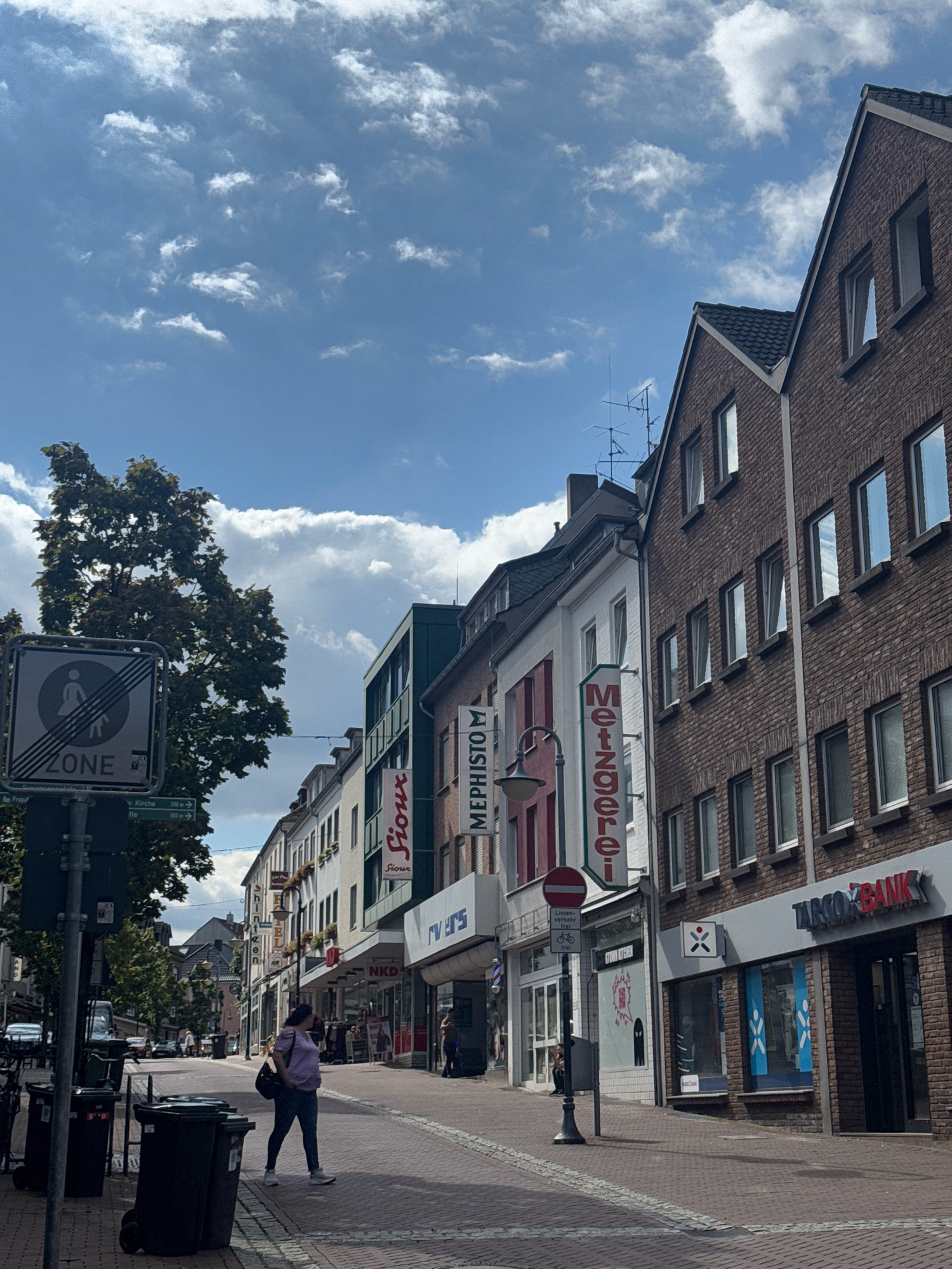 The image shows a street scene in Cleves, Germany. The street is paved with red bricks and lined with buildings on both sides. On the left side of the street, there is a tall green tree and a signpost with several signs, including a "ZONE" sign with a symbol of a person walking. Further down the street, there are shops with awnings and signs. On the right side of the street, there are brick buildings with several windows. One building has a sign that says "TARGO BANK".
A woman is walking on the sidewalk in the middle of the street, wearing a purple shirt and dark pants, carrying a dark bag over her shoulder. There are several trash bins on the left side of the sidewalk. The sky is blue with scattered white clouds. The overall impression is a sunny day in a European town.