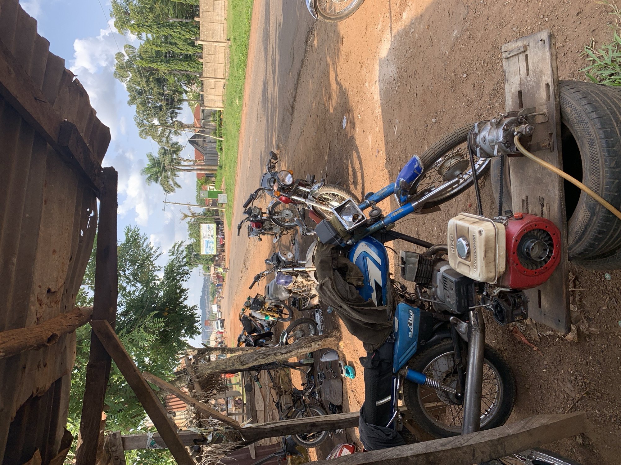 The image shows a collection of motorcycles parked on a dusty ground next to a dirt road. A corrugated metal roofed structure is partially visible in the foreground on the left. The motorcycles appear to be of various models and conditions, with some looking like they are being repaired or worked on. One motorcycle has a blue fuel tank and a red engine attached. There is also a spare tire visible, propped up on a wooden plank, with some mechanical equipment attached. In the background, there is a paved road with some vehicles and people, as well as buildings and trees. The sky is visible with some clouds, suggesting it is daytime. The overall impression is of a makeshift workshop or a parking area for motorcycles in a rural or semi-urban setting in Kabba, Nigeria. There are no explicit signs of people actively working on the motorcycles in this specific frame, but the presence of tools and equipment suggests ongoing activity.
