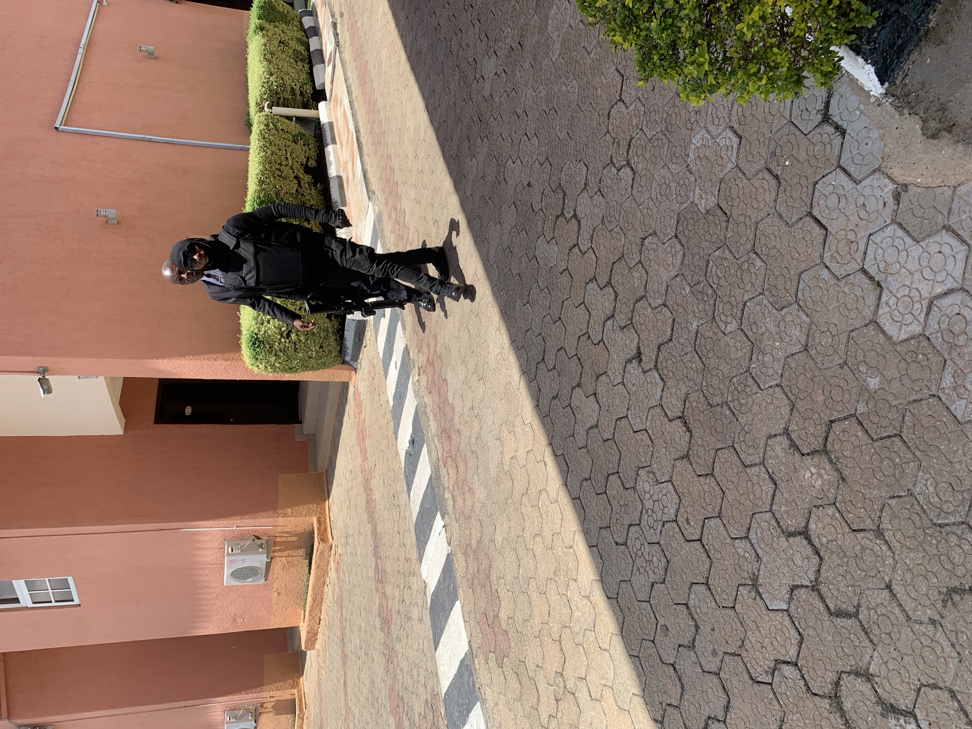 A man dressed in black tactical gear and a hood stands on a brick walkway in Gura Top, Nigeria. He holds a rifle. The walkway borders a building with a salmon-colored exterior and air conditioning units. A trimmed green hedge runs along the walkway. The walkway is paved with different colored bricks and features black and white striped markings.