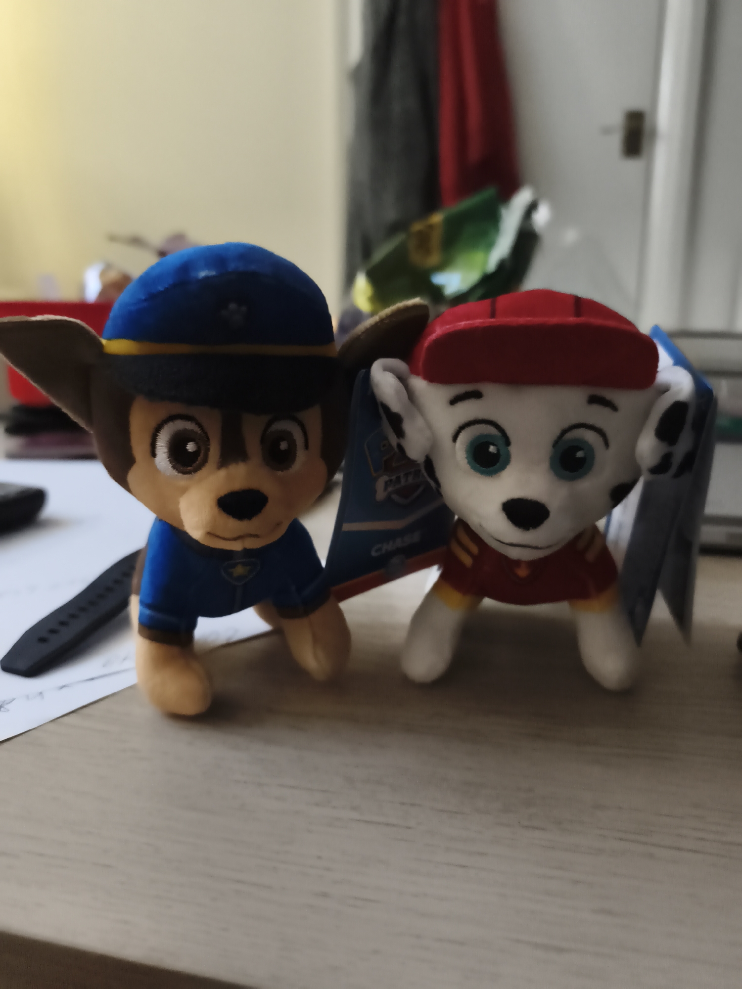 The image displays two plush toys, resembling characters from the animated series Paw Patrol, posed side-by-side on a light-colored wooden surface.
On the left is a plush toy of Chase, depicted as a German Shepherd wearing a blue police uniform with a gold badge and a blue police cap with a gold band. It has brown fur, embroidered brown eyes, and a small black nose, with light brown floppy ears.
To its right is a plush toy of Marshall, a white Dalmatian with black spots on its ears and around its eyes. Marshall wears a red fire uniform with yellow stripes on the sleeves and a matching red cap with a brim. It features large, bright blue embroidered eyes and a black nose.
Both plush toys are in a low, crawling-like pose. A product tag, partially visible between them, shows the text "PAW PATROL" and "CHASE." In the foreground, beneath the Chase plush, a black wristband or watch strap lies on a piece of white paper with some illegible handwritten text.
The background is blurred, indicating an indoor setting. Visible elements include light-colored walls or a door frame, a door with a handle, and some hanging fabric or clothing, including a red item and a green item further back. The general illumination suggests it is daytime. The scene is set indoors, within what appears to be a home in Huntingdonshire, United Kingdom.