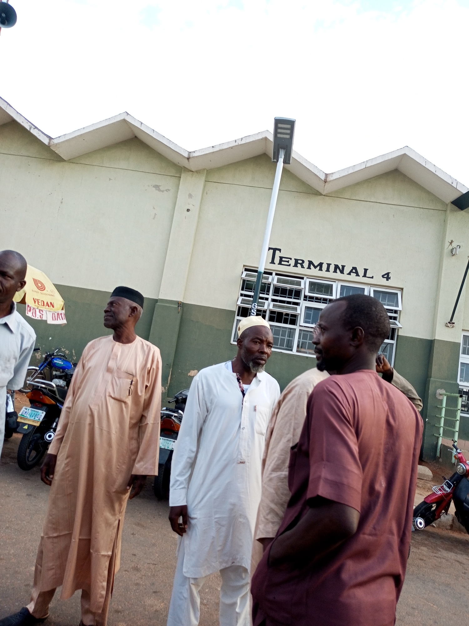The image depicts a street scene in Gombe, Nigeria, in front of a building labeled "TERMINAL 4". Several men are gathered, appearing to be in conversation. The building features a distinctive zigzag roofline and a series of windows. Various motorcycles are parked in the background. One man on the far left is wearing a light-colored shirt. A man in the center is wearing a tan robe and a black cap. To his right, another man is wearing a white robe and a white cap. The man in the foreground on the right is wearing a maroon robe. The overall environment suggests an outdoor waiting area or gathering point.