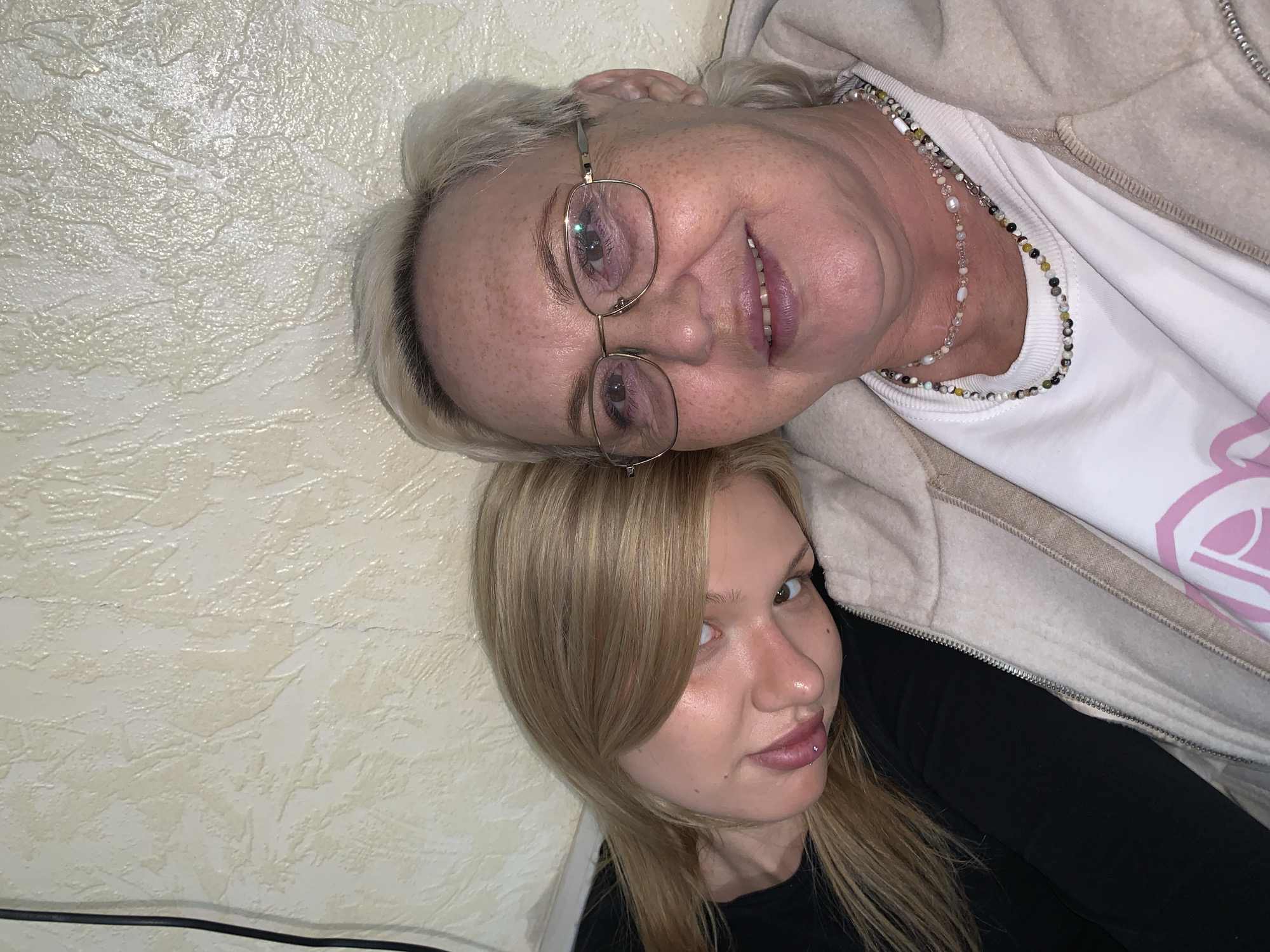 Two individuals are visible in the photograph. An older woman with short, gray hair, wearing glasses and a beaded necklace, smiles towards the camera. Below her, a younger woman with blonde hair poses, showing a lip piercing. Both individuals appear to be lying down, possibly on a textured, light-colored surface. It is impossible to determine the location as Chaikovsky, Russia with only the given visual information.