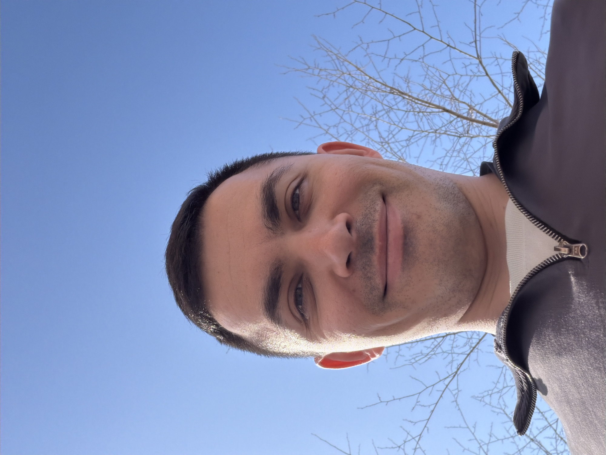 A close-up, low-angle selfie of a man against a clear blue sky with bare tree branches visible. The man is smiling slightly. He has short dark hair and is wearing a zippered dark-colored jacket with a white shirt underneath. The location is not discernible as Izhevsk, Russia.