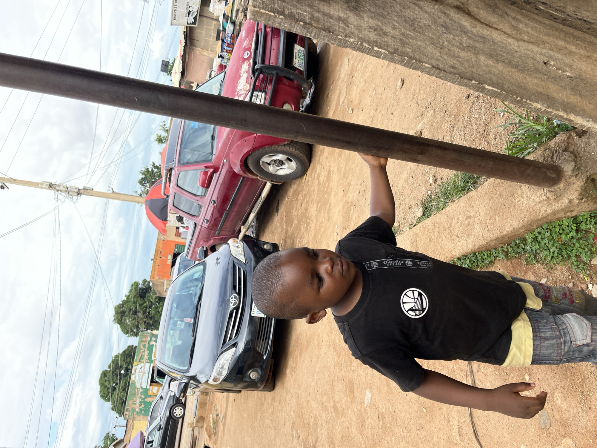 This image captures a street scene in Jos, Plateau State, Nigeria. In the foreground, a young boy with dark skin and a short haircut stands on a dirt path, looking downwards and slightly to his right. He wears a black t-shirt with a white circular Golden State Warriors logo, a light yellow undershirt, and faded blue jeans with cargo pockets and subtle designs. His right hand holds a vertical, dark metal pole anchored into a concrete curb that delineates the dirt path from an adjacent patch of green vegetation.
Behind the boy, a black Toyota Corolla sedan is parked on the dirt. Its Nigerian license plate is partially visible, indicating "PLATEAU" state. Further into the midground, a red Nissan Frontier pickup truck with a front bull bar is parked. Its Nigerian license plate clearly displays "PLATEAU" at the top, confirming the location.
The background features several single-story buildings under a cloudy, overcast sky. One building has a faded yellow-green facade with white decorative elements, while another appears reddish-orange. Utility poles and a network of overhead power lines crisscross the sky. To the far right, above the red truck, a partially visible sign reads "KINGS COLD STORE" with a phone number (0803878898). A red and black market umbrella is also visible above the red truck, alongside various goods displayed on the ground, suggesting a street vendor or market setup.