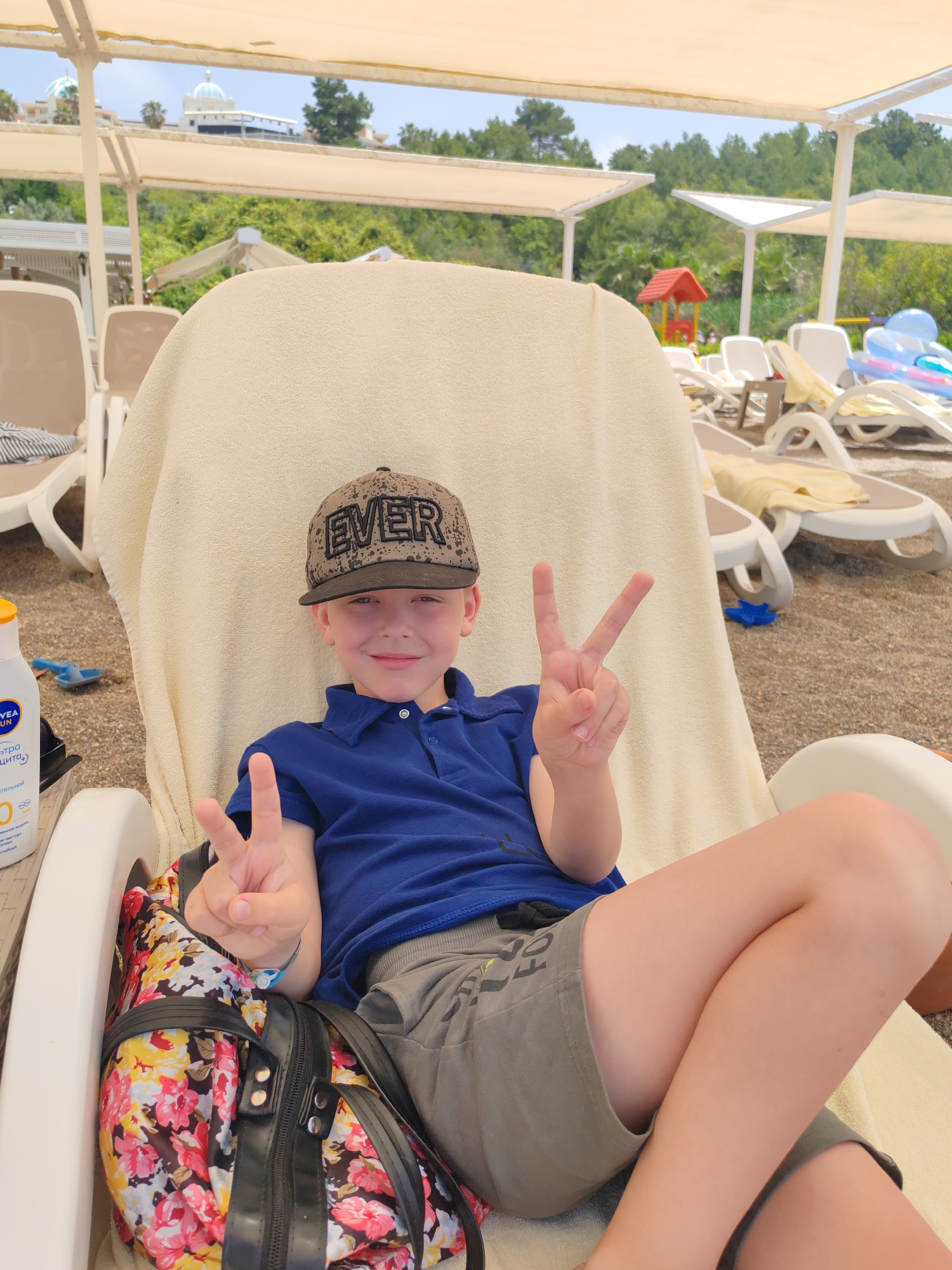 A fair-skinned young boy wearing a hat with the word "EVER" in black block letters, a navy blue polo shirt, and grey shorts is seated in a white plastic beach chair under a sunshade on a beach in Alanya, Turkey. He is holding up two fingers in a peace sign with each hand. A large floral-patterned bag is on the chair beside him. Behind him are more beach chairs, and a red play structure is visible in the background, with green trees and foliage dominating the background and a tall building visible beyond the trees. A bottle of Nivea sunblock is sitting on the chair to the left of the boy. The beach is covered in sand.