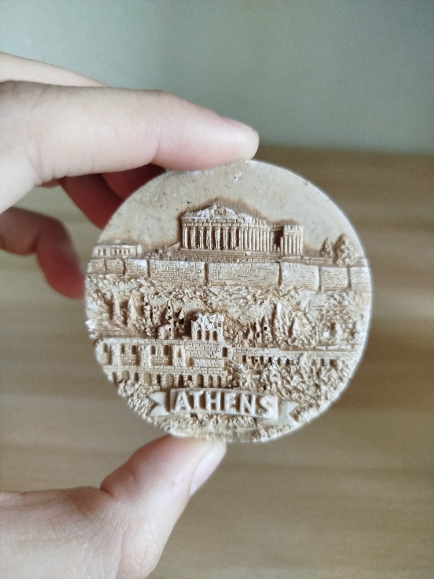 A hand is holding a round, beige souvenir of Athens, Greece. The souvenir depicts the Acropolis with the Parthenon at the top, a wall, trees, and other buildings. The word "ATHENS" is printed at the bottom of the souvenir. The background is out of focus and appears to be a wooden surface. The image does not show any location in the city of Varna, Bulgaria.