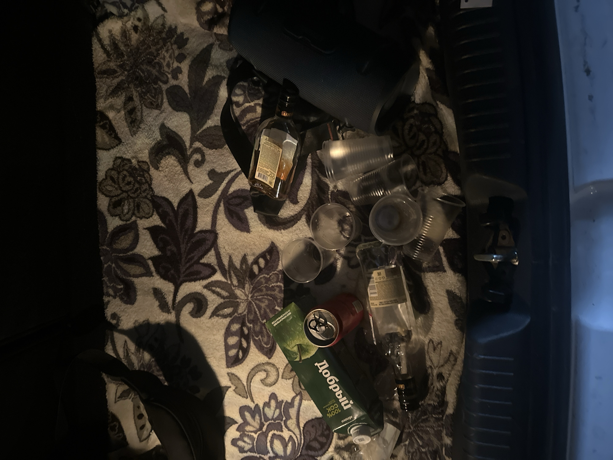 The image depicts a cluttered scene likely inside a vehicle. Scattered across a patterned blanket are an opened bottle of Oakheart rum, several empty plastic cups, an open can of what appears to be an energy drink, and a box of "Dobryy" juice. A portable Bluetooth speaker is also present. Given the presence of "Dobryy" juice, the location is likely in a Russian-speaking area, possibly Kazan, Russia. No people are visible.