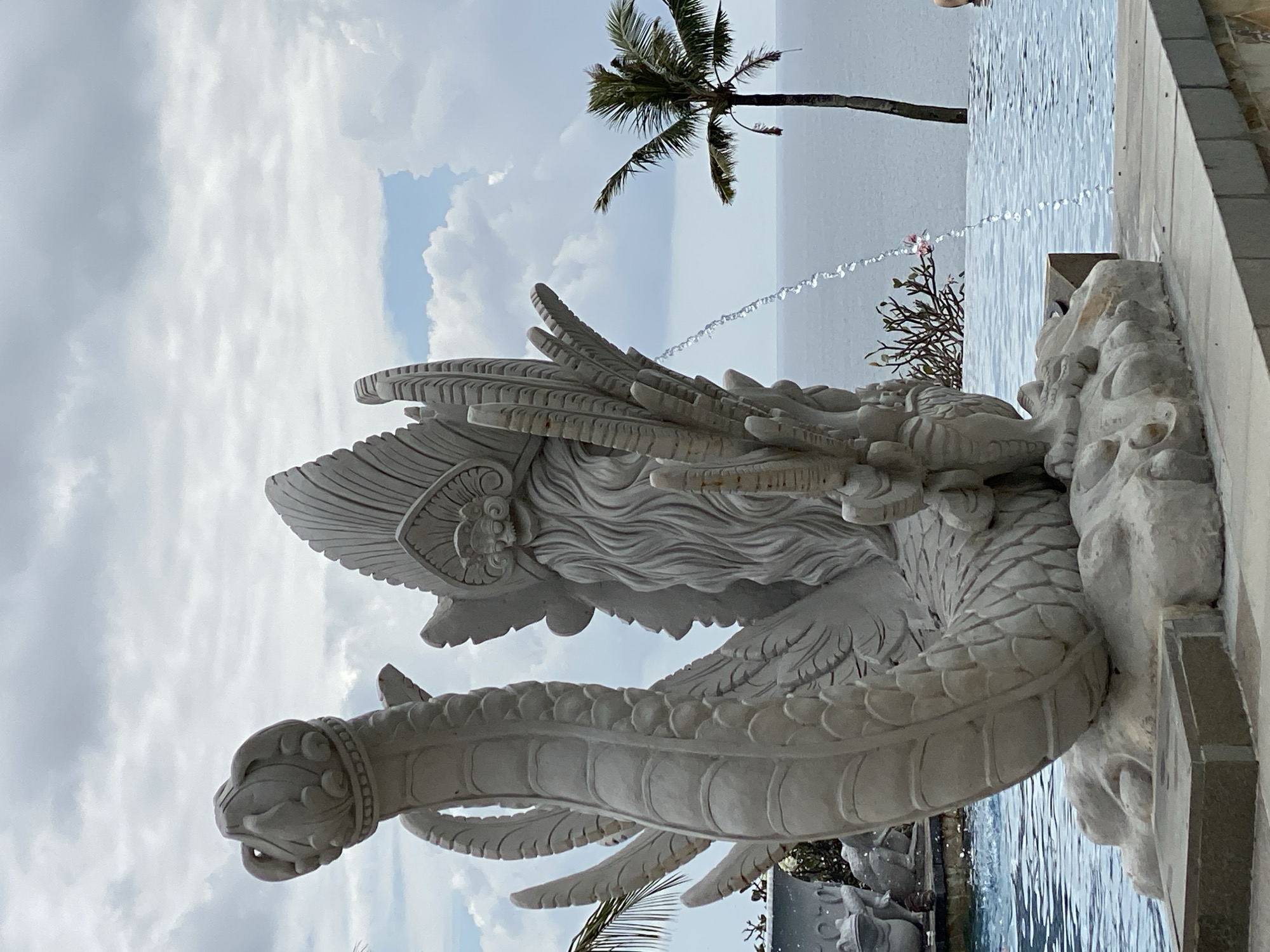 A stone statue of a mythological creature, possibly a Naga or a dragon, sits in the foreground of an infinity pool in Kuta Selatan, Indonesia. The statue is intricately carved, depicting scales, a headdress, and a long, flowing beard. Water streams from the statue into the pool, creating ripples. In the background, the ocean stretches to the horizon under a cloudy sky. A palm tree stands to the right of the statue, and a portion of a building can be seen to the left. The scene appears to be set at a resort or a hotel, given the presence of the pool and the tropical foliage. The lighting suggests it is daytime, with overcast conditions.