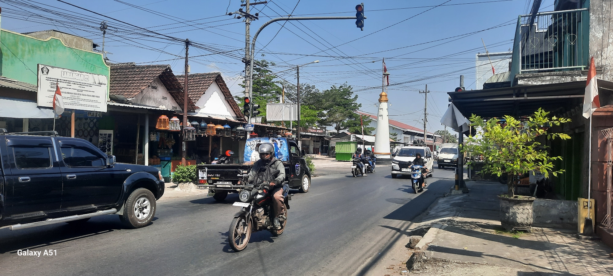 The image captures a street scene in Selopuro, Indonesia, on a bright, sunny day. The road is paved and shows signs of wear. On the left, a black pickup truck with tinted windows is parked or driving slowly. Further down the street, a motorcycle with a rider wearing a helmet and a backpack is moving forward. A black pickup truck with a graphic design on its side is also visible on the road, carrying what appears to be a load. Several other motorcycles and vehicles, including a white minivan, are present, indicating a moderate level of traffic.
The sides of the street are lined with buildings. On the left, a building with a green facade displays a sign with Indonesian text. Adjacent to it are shops with open fronts, one of which appears to be selling birdcages. Further along, a white tower-like monument stands at an intersection, topped with an Indonesian flag. Other buildings along the street have tiled roofs and visible electrical wires overhead. A small tree in a decorative pot sits on the sidewalk in the foreground on the right, casting shadows on the pavement. The sky is clear and blue.