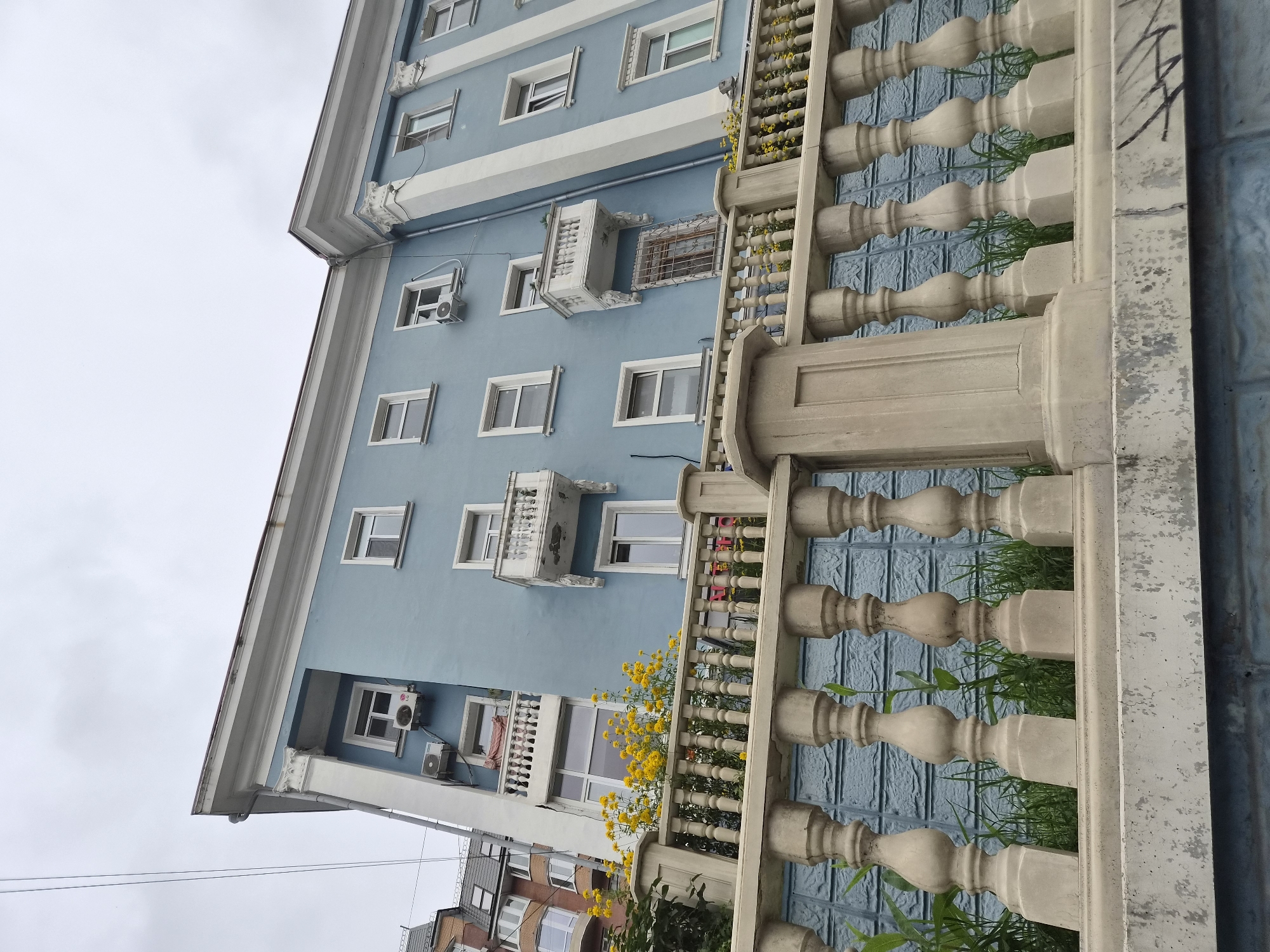 This is a low-angle, exterior, eye-level shot of a light blue apartment building facade in Vladivostok, Russia. In the foreground, a white stone balustrade with decorative spindles runs horizontally across the frame. Behind the balustrade, a textured blue wall is visible with green grass growing in front of it. Above the balustrade, yellow flowers are blooming on a section of the building. The building itself has multiple windows and balconies with white railings. There are air conditioning units mounted on the facade. To the left, a portion of another building with a multi-pitched roof and brown brick facade is visible. The sky above is overcast and gray. A black power line stretches across the upper left corner of the image. There are no people visible in the scene.