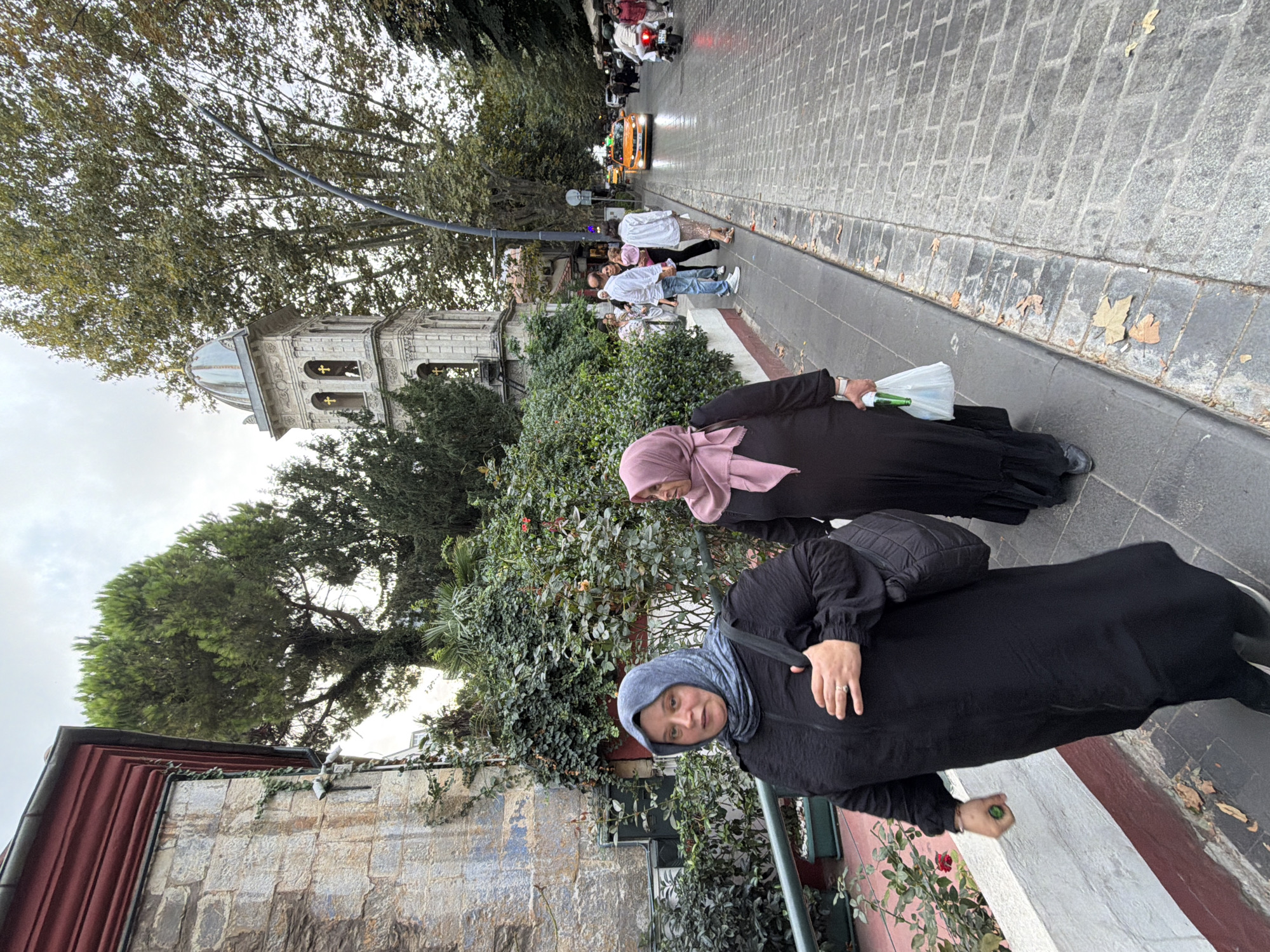 Two women wearing hijabs walk on a cobblestone sidewalk in Üsküdar, Turkey. The woman in the foreground wears a black abaya and a blue hijab, carrying a black bag over her left shoulder. The woman behind her wears a black abaya and a pink hijab. A bell tower of a building, possibly a church, stands to the left of the women, with trees and greenery surrounding it. In the background, a yellow taxi and a motorcycle are visible on the street, with other pedestrians walking along the sidewalk. The sky is overcast.