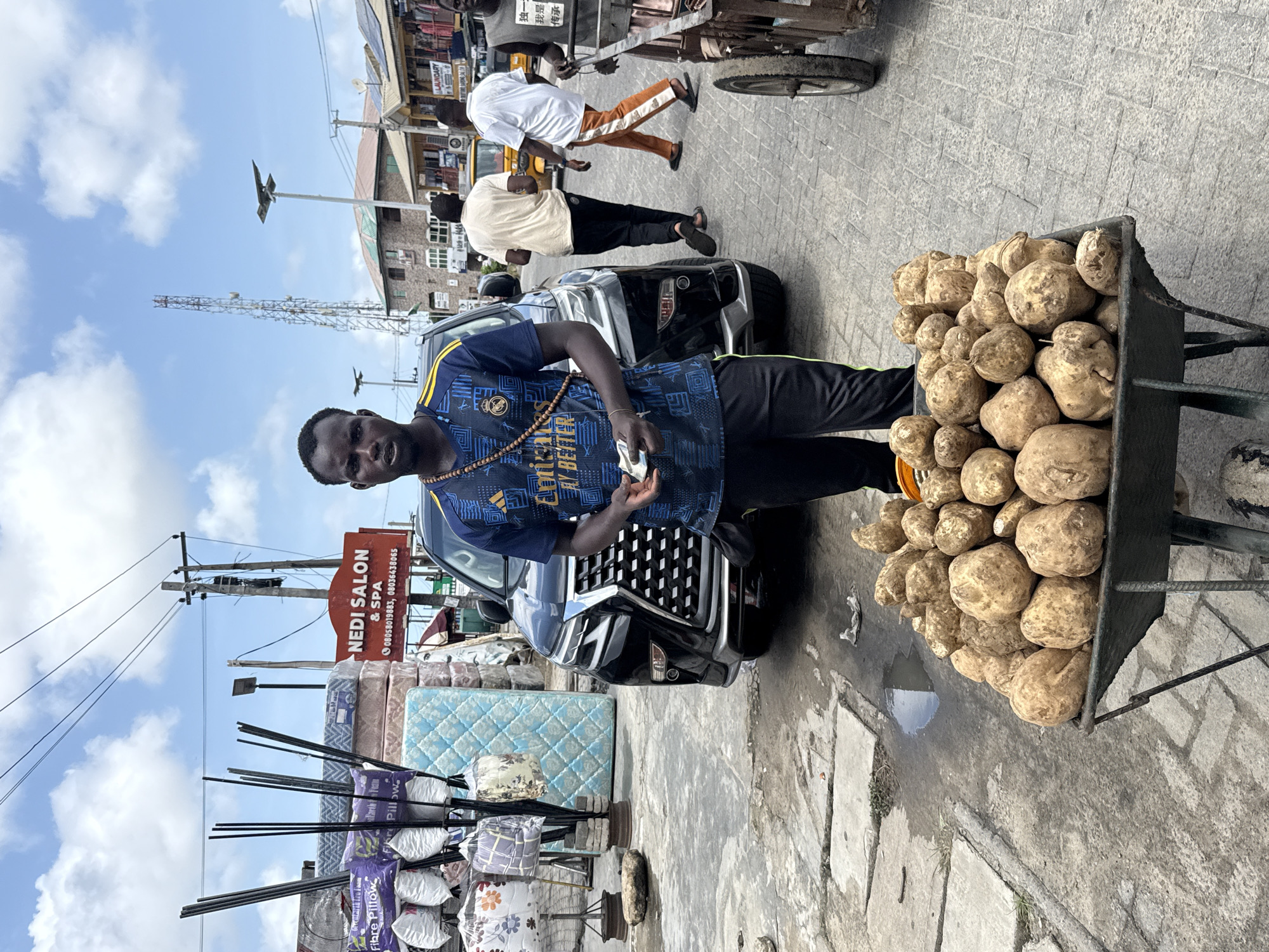 An eye-level, medium shot captures a man standing beside a wheelbarrow filled with yams, likely in Ado, Nigeria. He is clad in a dark blue t-shirt with light blue patterns and dark-colored pants. The yams are piled high in the wheelbarrow to his right. He holds a handful of folded currency in his left hand.
Behind the man, a black SUV is parked on the street, and pedestrians walk in the background. A sign reading "NEDI SALON & SPA" is visible, as are mattresses and pillows displayed for sale. Utility poles with electrical wires stretch overhead. The street and sidewalk are made of interlocking pavers, showing wear and some debris. The sky is partly cloudy.