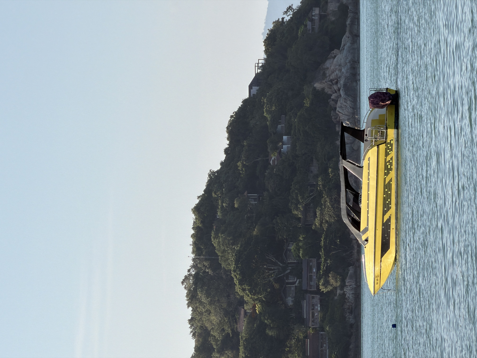 A vibrant yellow speed boat with a black canopy is anchored in the calm, turquoise waters off the coast of Ko Samui District, Thailand. The boat features a sleek design with black accents and a textured honeycomb pattern along its hull. A dark red cover is draped over the stern. In the background, a lush green, tree-covered hillside rises from the water's edge. Houses are nestled within the foliage on the slopes, some appearing to be modern structures with dark roofs, while others are more traditional bungalows. Rocky outcrops are visible amidst the trees. The sky above is a clear, pale blue. The scene depicts a serene tropical setting.