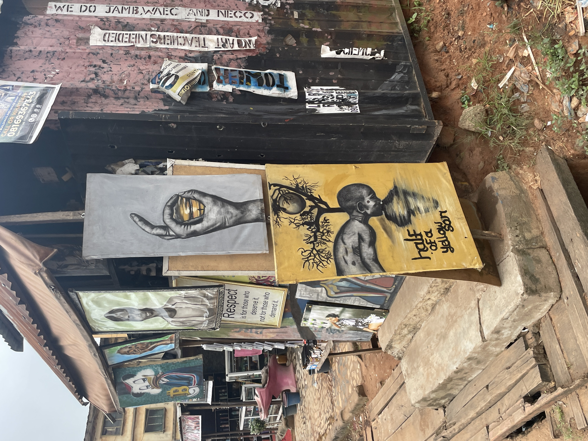 The image depicts an outdoor art stall located in Nkpor, Nigeria. A corrugated metal roof extends over a display of paintings, some of which are hung on the wall of a dark, possibly shipping container-like structure. Several artworks are displayed prominently, including a painting of a hand holding a coin, a portrait with the text "Respect," and a yellow painting with the text "half of a yellow sun." Other paintings, some abstract and others featuring portraits, are also visible. In the background, shelves stocked with various items are seen within a building. Wooden planks form a pathway and steps leading up to the stall. The ground around the stall is dirt with some patches of grass and scattered debris. There are no people visible in the immediate scene. The environment appears to be a street or market area, consistent with a commercial district in Nkpor.
