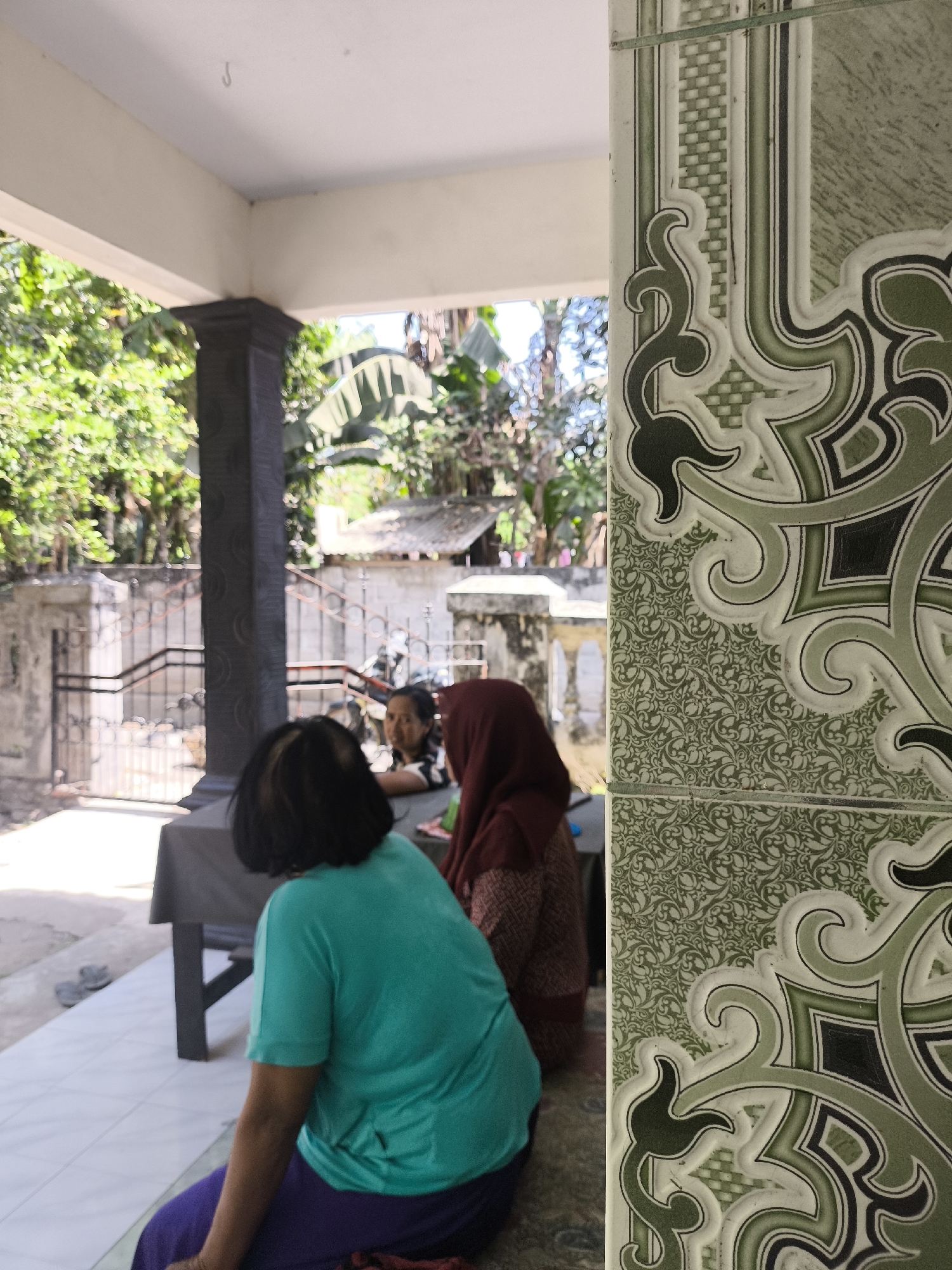 The image depicts an outdoor scene in what appears to be a residential area in Grati, Indonesia. In the foreground, a person wearing a teal shirt and purple pants is seated on the floor, facing away from the camera. To the right, a portion of an ornate, green tiled pillar is visible. In the background, partially obscured by the pillar and the seated person, two other individuals are visible. One person, wearing a maroon headscarf and patterned clothing, is partially seated, and another person with dark hair is looking towards the camera. Behind them, there is a table, a dark-colored pillar, and a fence. Lush green foliage, including banana trees, suggests a tropical or sub-tropical environment. The overall impression is a casual gathering in an outdoor, possibly domestic, setting.