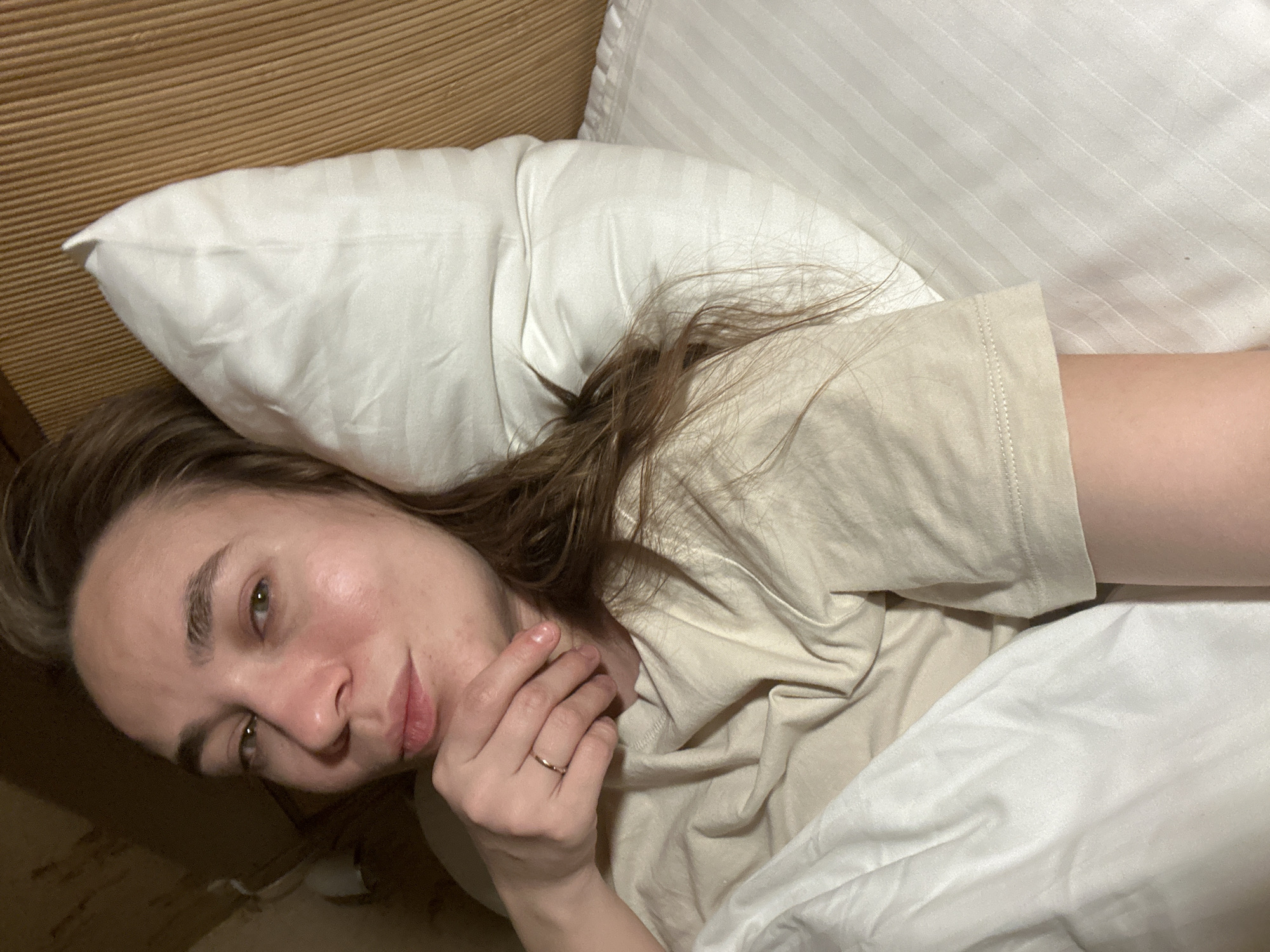 A woman lies in a bed with white pillows and a white comforter. She is wearing a light tan t-shirt and a gold ring on her left ring finger. She has brown hair and green eyes. The headboard is brown. The location cannot be confirmed as Yekaterinburg, Russia.